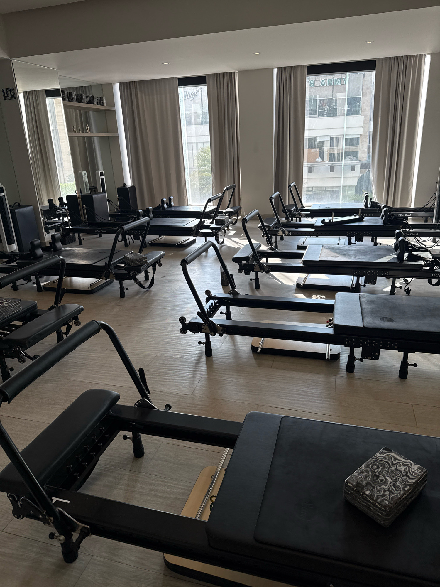 The image depicts an indoor fitness studio in San Bernardino Tlaxcalancingo, Mexico. The room is filled with several black Pilates reformers arranged in rows on a light-colored wooden floor. Large windows with sheer, light-colored curtains cover the walls, allowing natural light to illuminate the space. Through the windows, buildings and sky are visible, indicating an urban setting. The studio appears to be empty, with no individuals present. The overall atmosphere is clean and organized, suggestive of a professional fitness facility.