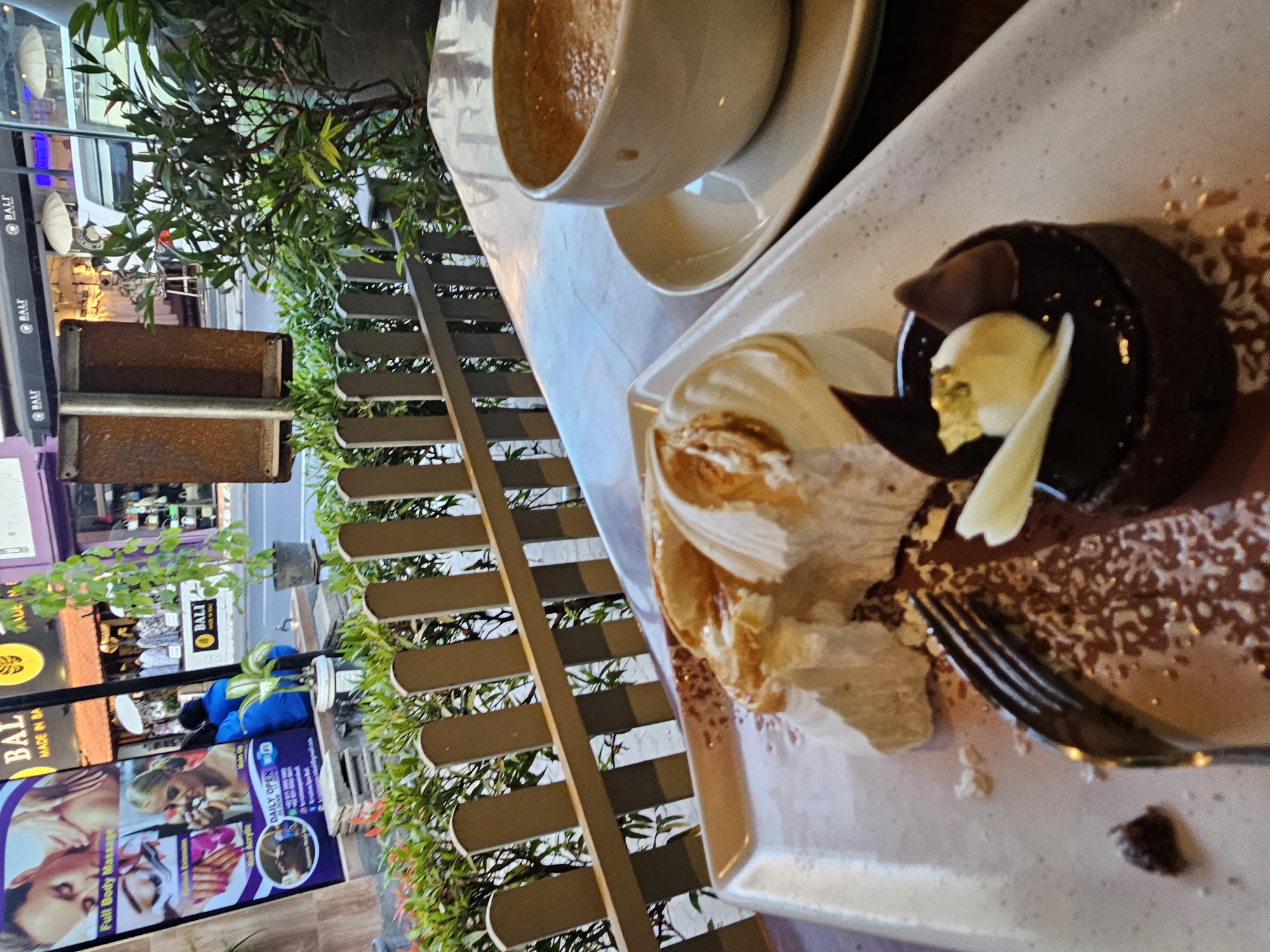 The image shows a table setting at an outdoor cafe with a view of a street in Denpasar, Indonesia. In the foreground, a plate holds a partially eaten meringue dessert with caramel sauce and a small chocolate cake. A fork rests on the plate. To the right, a cup of coffee sits on a saucer.
Behind the table, a wooden fence separates the cafe from the street. A lush green plant is visible next to the fence. Further back, a street scene unfolds with shops and people. A sign on the left advertises "Full Body Massage," "Eyelash Extension," and "Nail Acrylic." A white van is parked on the street, and several people are walking or standing around. The overall atmosphere suggests a casual dining experience in a bustling urban environment.