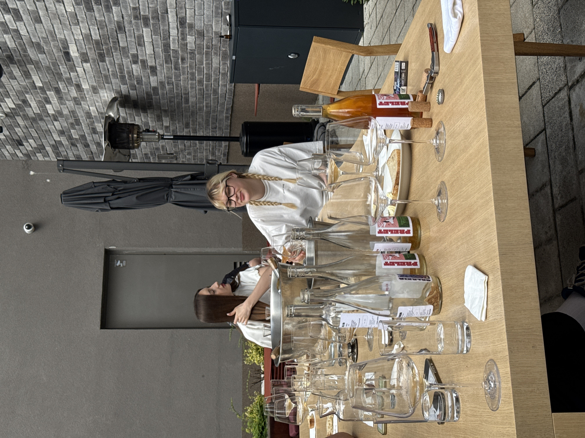 The image shows an outdoor setting, likely in Belgrade, Serbia, featuring a long wooden table set for a wine tasting. The table is covered with numerous empty wine glasses and several empty bottles labeled "PRELET". Two women are present; one with blonde hair in braids wearing glasses and a white shirt, and another with long brown hair, her back turned. They are positioned near the table, suggesting they were part of the tasting event. An ice bucket is on the table as well. The background shows a gray wall and a partially visible door, along with an outdoor patio heater and an umbrella. In the upper portion of the image, brick walls and a portion of a building are visible. On the table, aside from the glasses and bottles, are a wine opener, corks, and a few packs of cigarettes.
