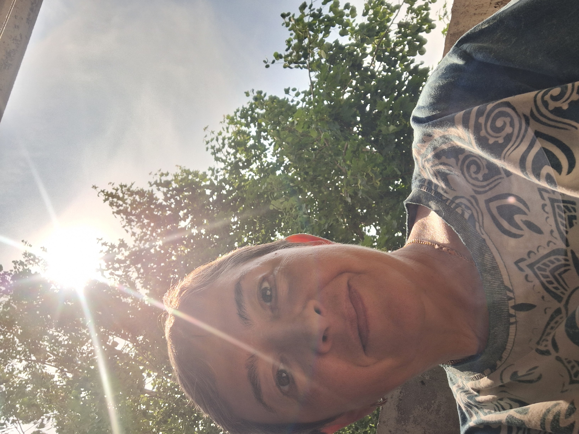 A close-up, selfie-style shot captures a person with short, dark hair and brown eyes, looking towards the camera with a slight smile. The sun is bright, creating a lens flare effect and casting light across the face. The person is wearing a t-shirt with a blue and white patterned design, a gold chain necklace, and small hoop earrings. In the background, lush green foliage from trees is visible, with a bright sky peeking through the branches. A concrete wall is also present in the lower left corner. The image appears to have been taken outdoors, possibly in a garden or park setting. It is not possible to determine the specific location within the city of Gatchina, Russia from the visual information provided.