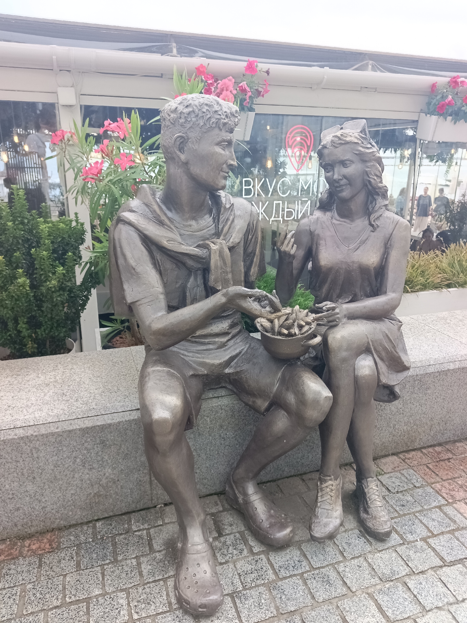 A bronze sculpture depicting a young couple sitting on a stone bench is located in the city of Sochi, Russia. The man, on the left, wears a t-shirt, shorts, and clogs, with a scarf draped around his neck. He holds a bowl filled with food, offering it to the woman. The woman, on the right, is dressed in a simple dress and wears sneakers. She is looking at the man and appears to be reaching for food from the bowl. Both figures have a relaxed posture, suggesting a casual, intimate moment.
Behind them, large windows reflect the surroundings, and signage on the glass reads "ВКУС МОЛОДЫМ" (for the young). In the background, lush green bushes and pink flowers in planters add a touch of nature to the urban setting. The ground is paved with cobblestones. The overall scene captures a slice of everyday life, immortalized in bronze.