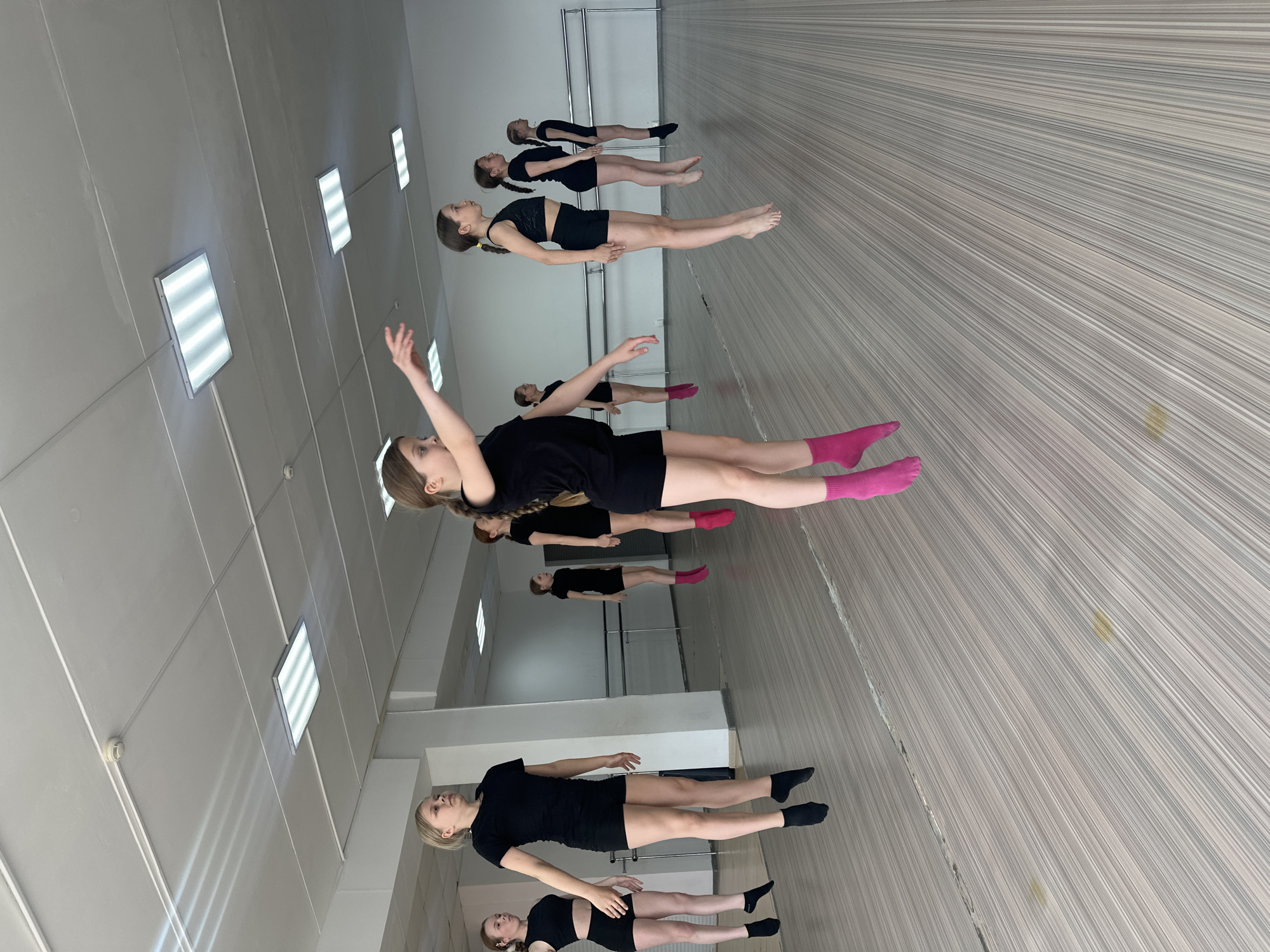 The image depicts a dance studio in what is likely Votkinsk, Russia, given the context provided. The studio features a wall-sized mirror reflecting a line of young female dancers. The dancers, dressed in black dance attire and socks of varying colors (black, pink), are engaged in a floor exercise. They are positioned horizontally with arms extended. The studio has a polished, striped floor and a barre along the mirrored wall. The ceiling is lit by recessed fluorescent lights. The overall impression is a professional and functional space for dance training.