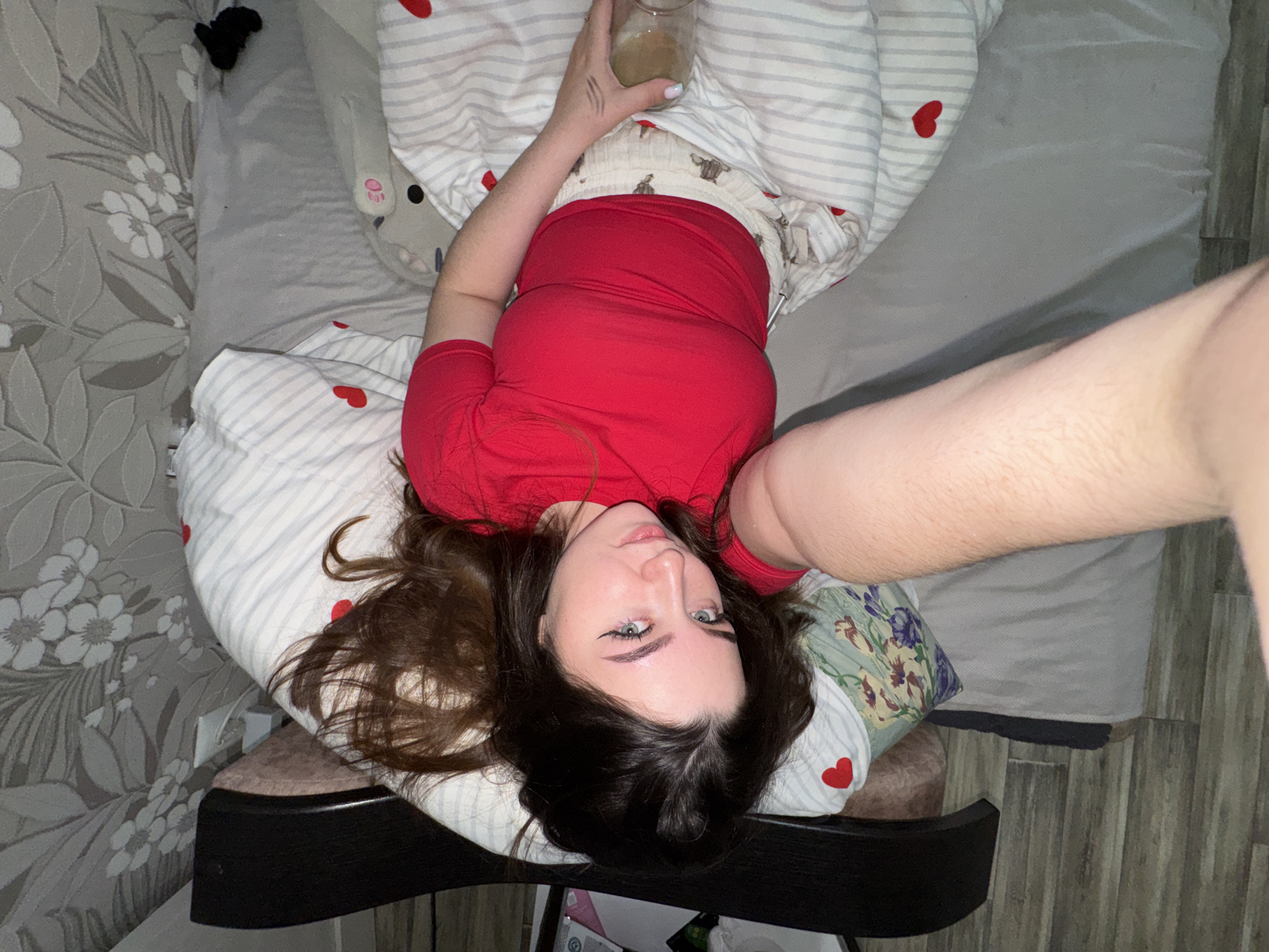 An overhead shot captures a young woman lying on her back, seemingly taking a selfie. She has long dark hair and light-colored eyes, accentuated by dark eyeliner and defined eyebrows. She is wearing a red t-shirt. Her right hand holds a glass or jar containing a light-colored liquid, and three short, parallel lines are drawn on the back of her hand. Her left arm is extended towards the camera, suggesting she is holding the device taking the photo.
She is lying on a bed covered with white bedding featuring thin grey stripes and small red heart patterns. A decorative pillow with purple, green, and white floral patterns is visible near her head. Another plush toy, grey with a pink paw print and black dots, is partially visible near her right shoulder.
The room's interior suggests a bedroom setting. The wall to her left is adorned with grey wallpaper featuring a pattern of white flowers and leaves. A dark-colored piece of furniture, possibly a stool or chair, is positioned near her head. The floor in the foreground and to the right appears to be made of wood or laminate. The lighting in the room is soft and ambient. The woman's expression is neutral as she looks directly at the camera. The setting is indoors in Gamovo, Russia.