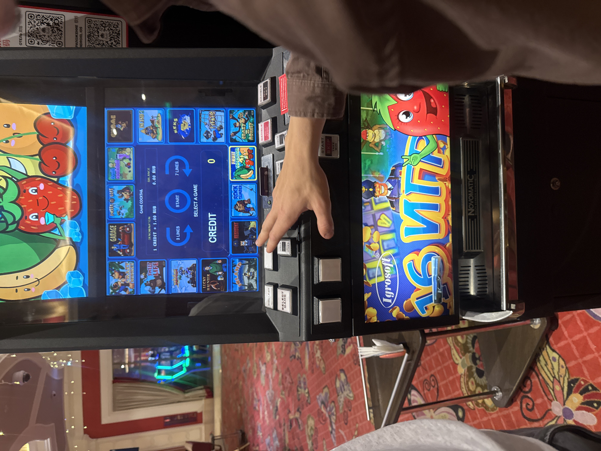 The image shows a person's hand interacting with a slot machine in what appears to be a casino or entertainment venue. The slot machine has a bright, colorful display featuring cartoon characters and game titles such as "Crazy Monkey," "Sweet Life," and "Fruit Cocktail." The denomination is set to "1 CREDIT = 1.00 RUB," and the balance is "0.00 RUB." The person's hand is positioned over the buttons, suggesting they are playing the game. In the background, through a glass partition, other slot machines with neon lights are visible, along with a red carpet with intricate patterns. The environment suggests an indoor entertainment setting. No specific details about the city of Shkotovo, Russia, are present within the image itself.