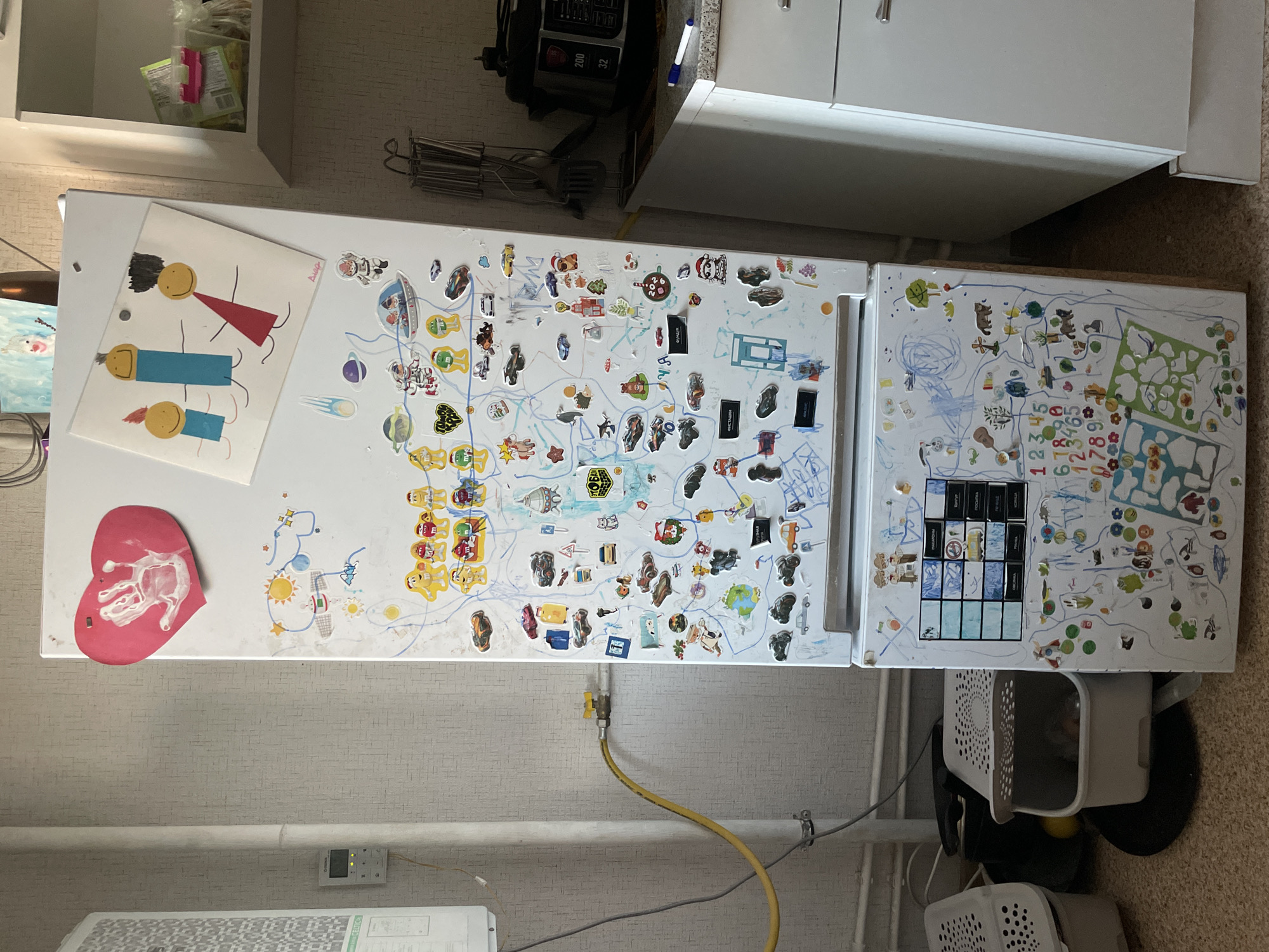 The image shows a close-up view of a refrigerator door covered in various stickers and drawings. The refrigerator is white and appears to be in a kitchen. To the left of the refrigerator is a wall with a textured pattern and a gas pipe with a yellow hose. Below the refrigerator are several white storage bins. The refrigerator door itself is decorated with children's artwork, including a handprint on a red heart sticker, drawings of stick figures, planets, spaceships, cars, animals, and numbers. Some of the stickers are also cartoon characters. The overall impression is of a lived-in space, likely a home, with a focus on children's activities and decorations. The city of Laishevo, Russia is not depicted in the image, nor is there any information that would suggest the location to be Laishevo.