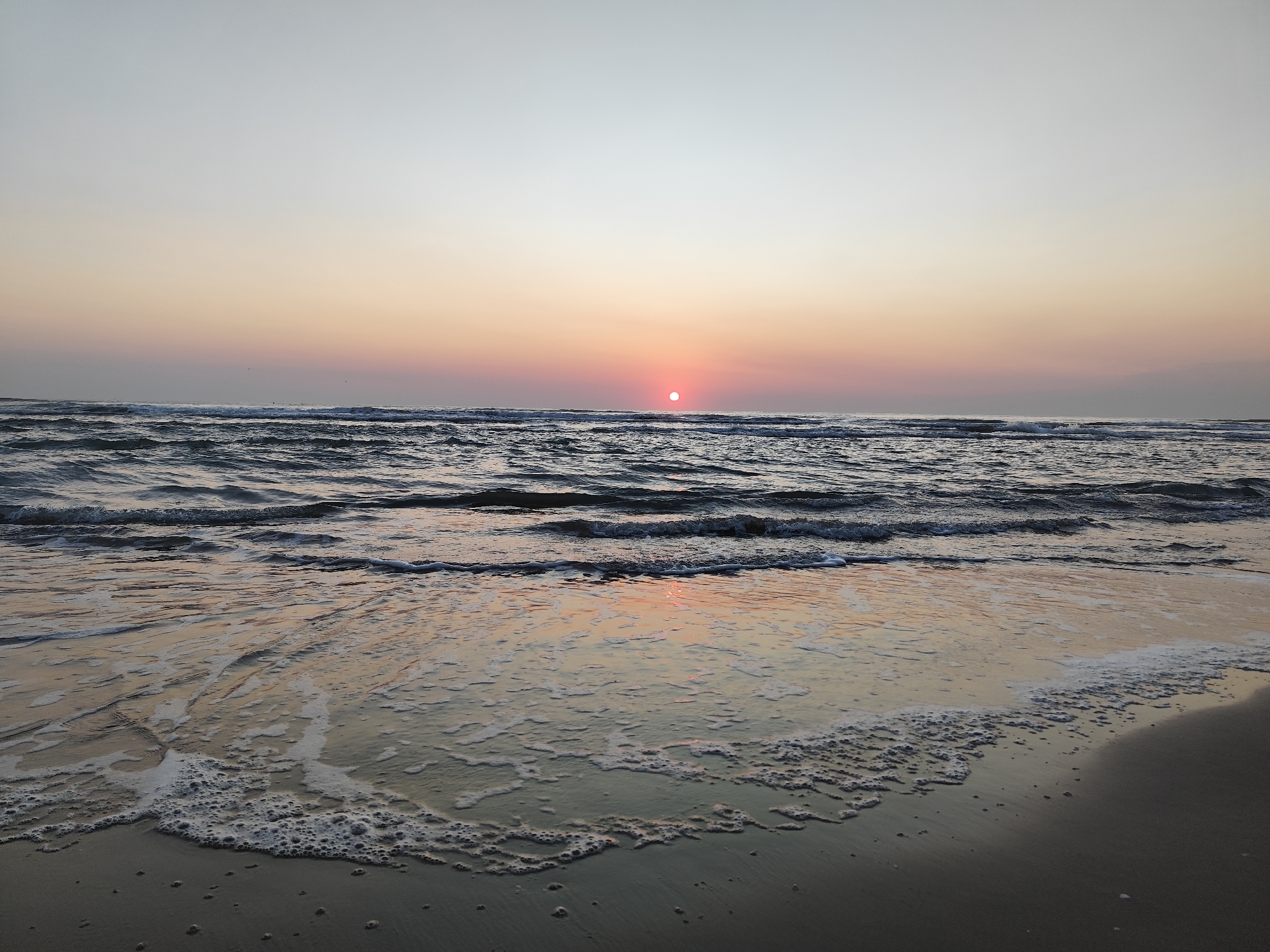 A tranquil beach scene at sunset in Rimini, Italy, presents a serene view of the Adriatic Sea. The sun, a small, bright red orb, is positioned just above the horizon, casting a warm, soft glow across the sky. The sky transitions from a pale blue at the top to shades of pink and orange closer to the horizon, indicating the fading light of day. Gentle waves roll onto the sandy shore, creating a thin line of white foam that stretches across the wet sand. The water is a deep blue-gray, reflecting the colors of the sunset. The sand is dark and wet where the waves have receded, with a few small ripples and scattered dark specks. The overall atmosphere is one of peacefulness and natural beauty, characteristic of a Rimini sunset. There are no people or man-made objects visible in the frame, emphasizing the natural elements of the scene.