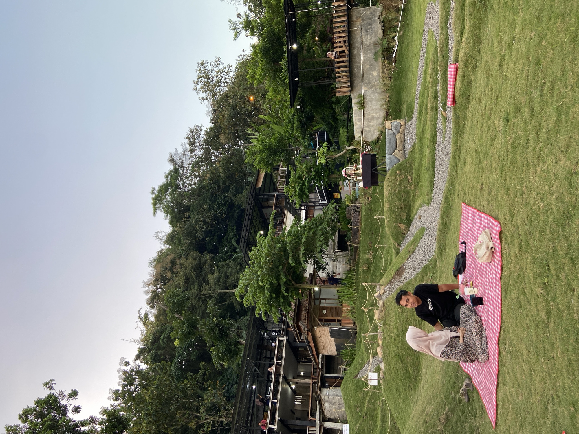 In Kesiman, Indonesia, a couple is picnicking on a red and white checkered blanket spread across a grassy hill. The man, wearing a black t-shirt, sits with his legs crossed, facing the woman. The woman, dressed in a floral patterned dress and a light pink hijab, is seated opposite him, also with her legs crossed. They appear to be in conversation, looking at each other. Scattered on the blanket are a couple of cups, a mobile phone, and a beige handbag. To their left, a pair of sandals rests on the grass.
The background reveals a multi-tiered establishment with wooden balconies and railings, built into a lush, green hillside. People can be seen on the balconies of the structure. Trees and vegetation cover the slope, with some outdoor lighting visible. A pathway made of gravel winds through the manicured lawn. In the mid-ground, a black table is set up with what appears to be decorative items, possibly for a romantic setup. The overall scene suggests a serene and scenic dining or recreational area. The sky is clear and a light blue, indicating it is likely late afternoon or early evening.