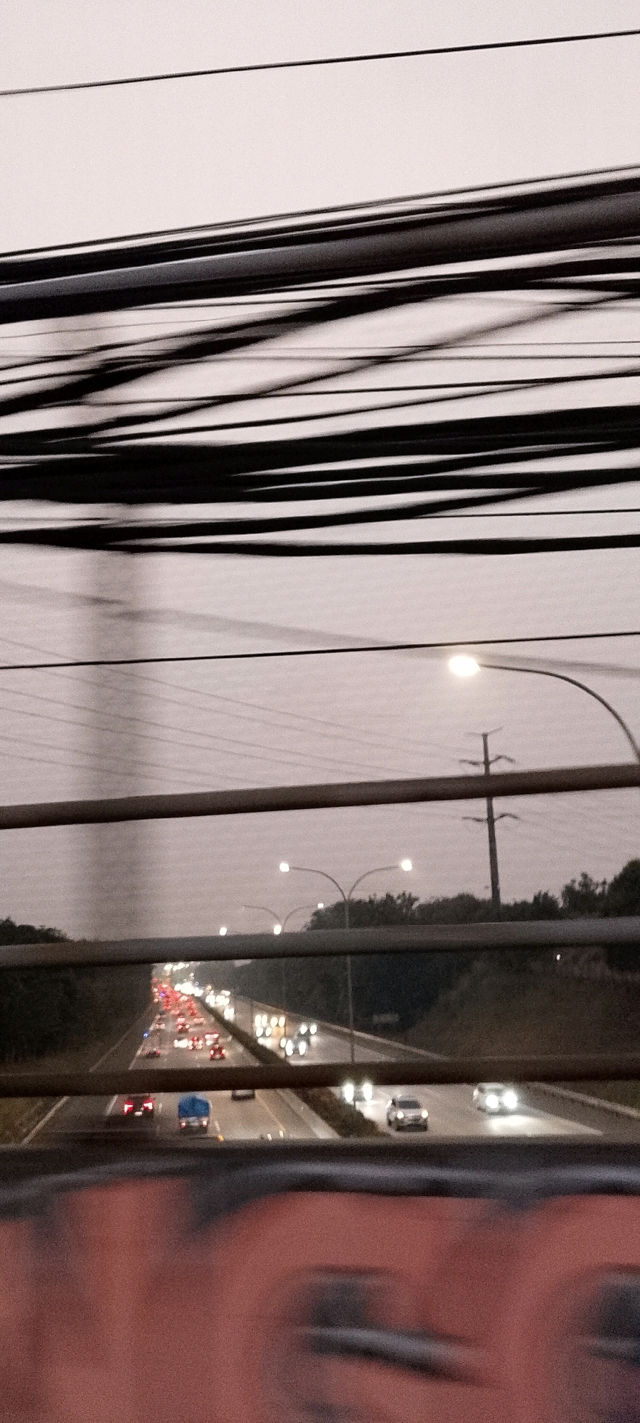 The image depicts a view from a pedestrian bridge overlooking a highway in Depok, Indonesia, likely during dusk or dawn given the diffused light and illuminated vehicles. Multiple parallel wires crisscross the upper portion of the frame, obscuring a portion of the sky. Below the wires, a highway stretches into the distance, with lanes filled with vehicles. Headlights and taillights of cars and trucks are visible, indicating traffic flow in both directions. Streetlights on poles are intermittently positioned along the highway's edge, casting pools of light. The foreground shows a blurred section of what appears to be the railing of the pedestrian bridge, with a blurry reddish-orange object below it, possibly a person's clothing or a printed image. The overall atmosphere is muted and somewhat hazy, characteristic of low light conditions and possibly some atmospheric moisture.