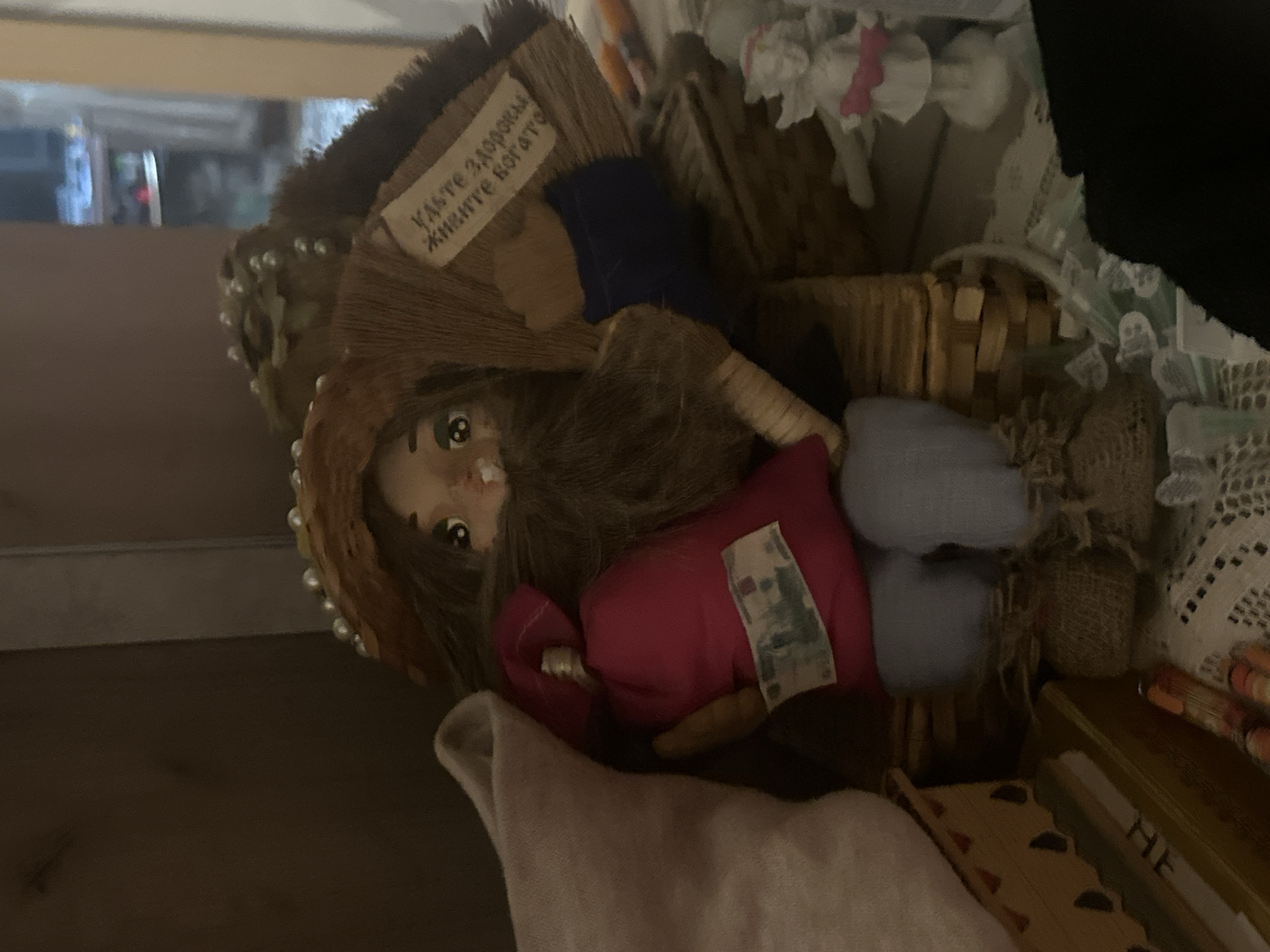 The image shows a doll lying on its side inside a wicker basket filled with other items. The doll has brown hair and wears a brown and blue outfit with a red cushion next to it. There is a small tag that reads “Касте здоровья живите богато”. Based on the Cyrillic script of the sign on the doll’s outfit, it is highly probable that the location is in Moscow, Russia, where Russian is the prevalent language.