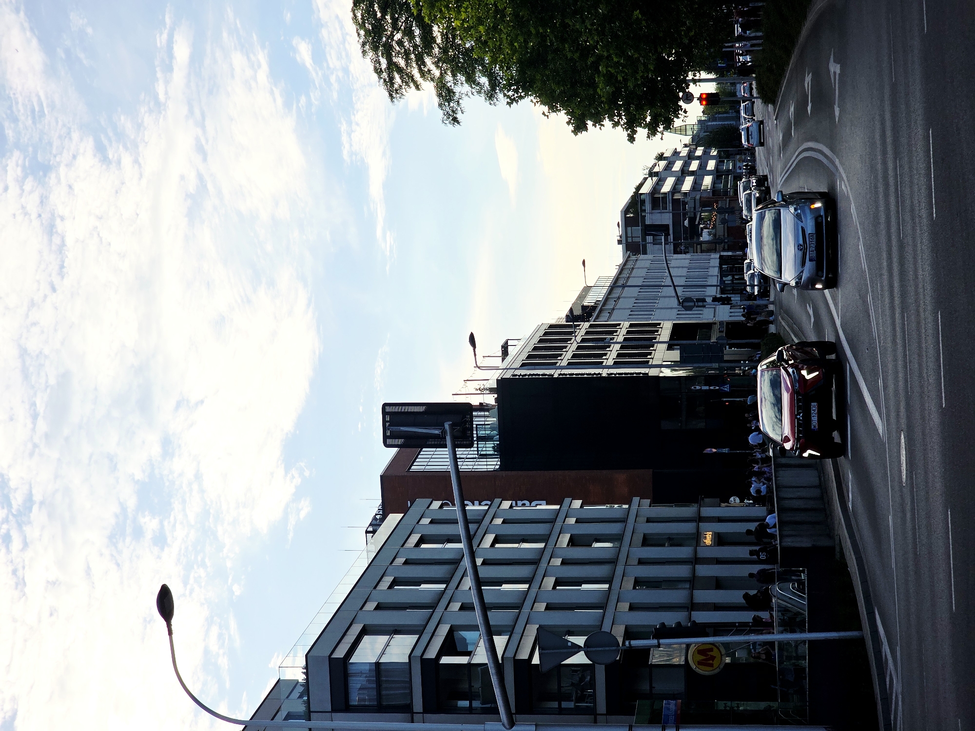 A street scene in Warsaw, Poland, shows a multi-lane road in the foreground with several cars driving. A red SUV is in the left lane, and a silver sedan is in the right lane. The road is bordered by modern buildings on the left and right. The buildings on the left are tall and grey with many windows, and a street lamp arches over the road. The buildings on the right are also modern, with some trees visible on the far right. The sky above is a mix of blue and white clouds. The image captures a busy urban environment in Warsaw.