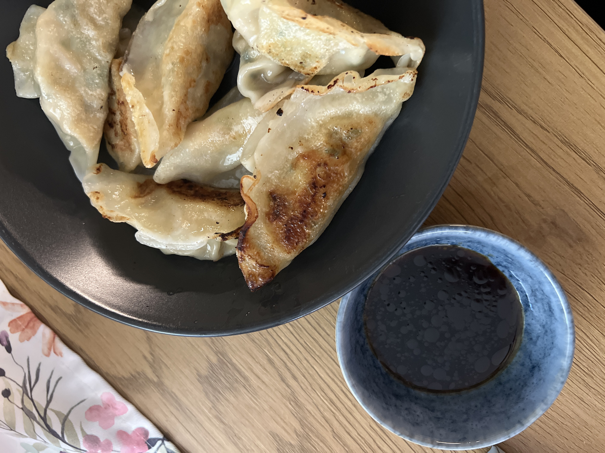 A high-angle shot presents a plate of pan-fried dumplings with a small bowl of dipping sauce, set on a wooden table. The dumplings are golden brown and appear to be potstickers. The sauce is a dark color, likely soy sauce or a mixture thereof. Part of a floral-patterned napkin is visible in the lower left corner. The scene is lit naturally. There is no indication of a location in Sofia, Bulgaria.