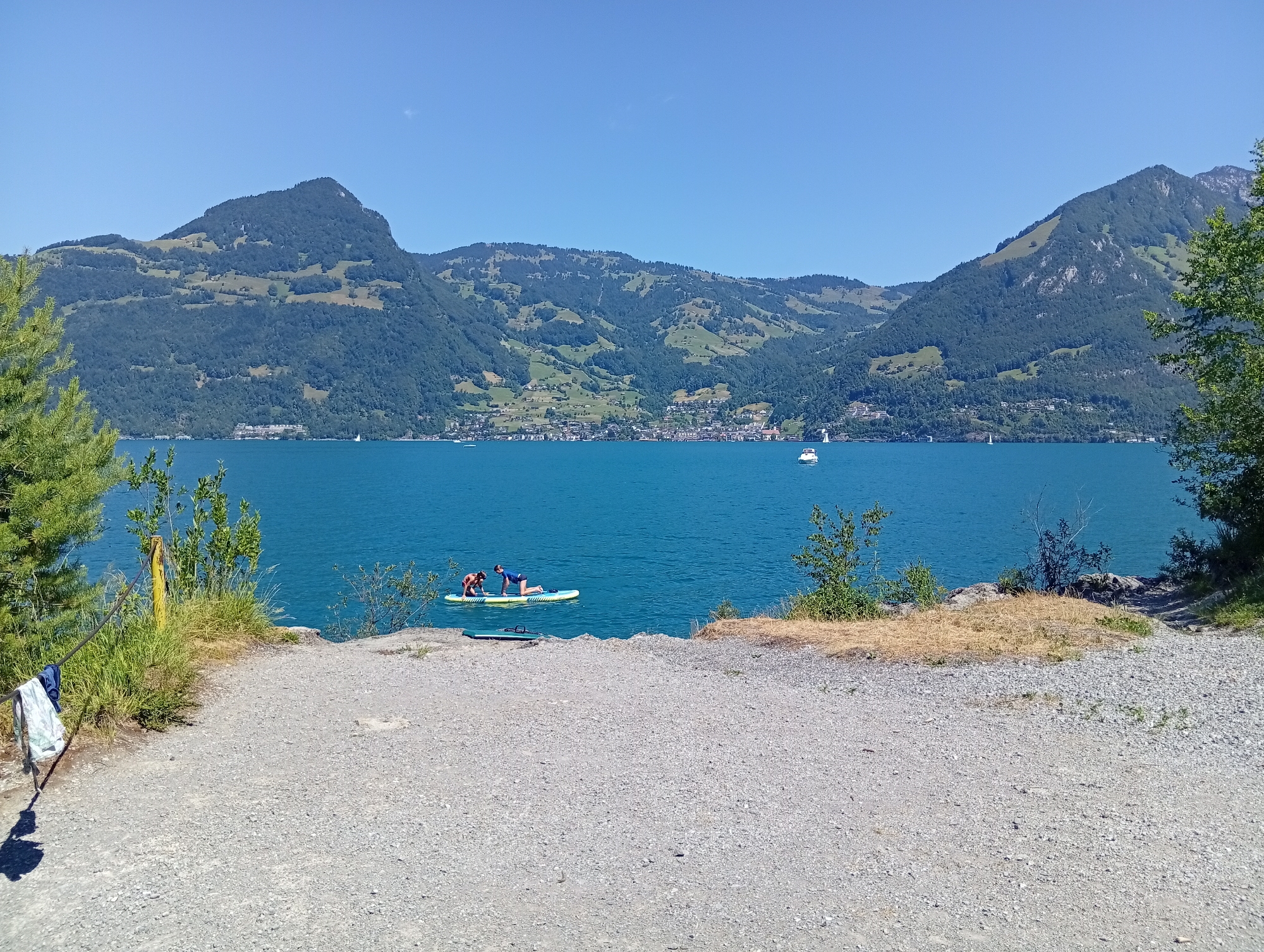 An eye-level, long shot captures a picturesque landscape on a clear, sunny day, with the foreground dominated by a gravelly path leading to a rocky edge overlooking a vibrant turquoise lake. Two individuals are seen on a paddleboard in the middle ground of the lake, one kneeling and the other in a push-up position. The lake itself is dotted with a few sailboats in the distance, and its far shore is lined with a town nestled at the base of lush, green mountains. These mountains, characteristic of Emmetten, Switzerland, display terraced fields and dense forests under a bright blue sky. To the left of the path, a post with a wire fence is adorned with a white and blue garment hanging to dry. The scene evokes a sense of tranquil outdoor activity.