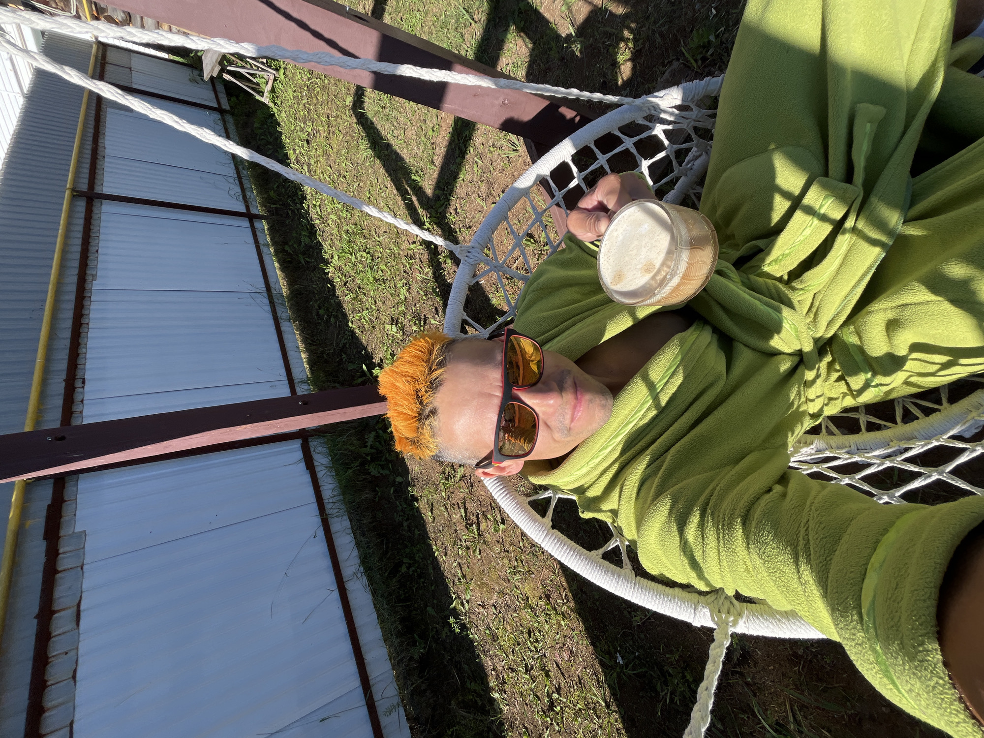 In Chaikovsky, Russia, a person with bright orange hair and sunglasses is sitting in a white macrame hanging chair outdoors. They are wearing a green robe and holding a glass of coffee with foam. The background shows a corrugated metal fence with a yellow pipe running along the top, green grass, and the shadow of the chair and person cast on the ground from the sunlight. A wooden structure supports the chair.