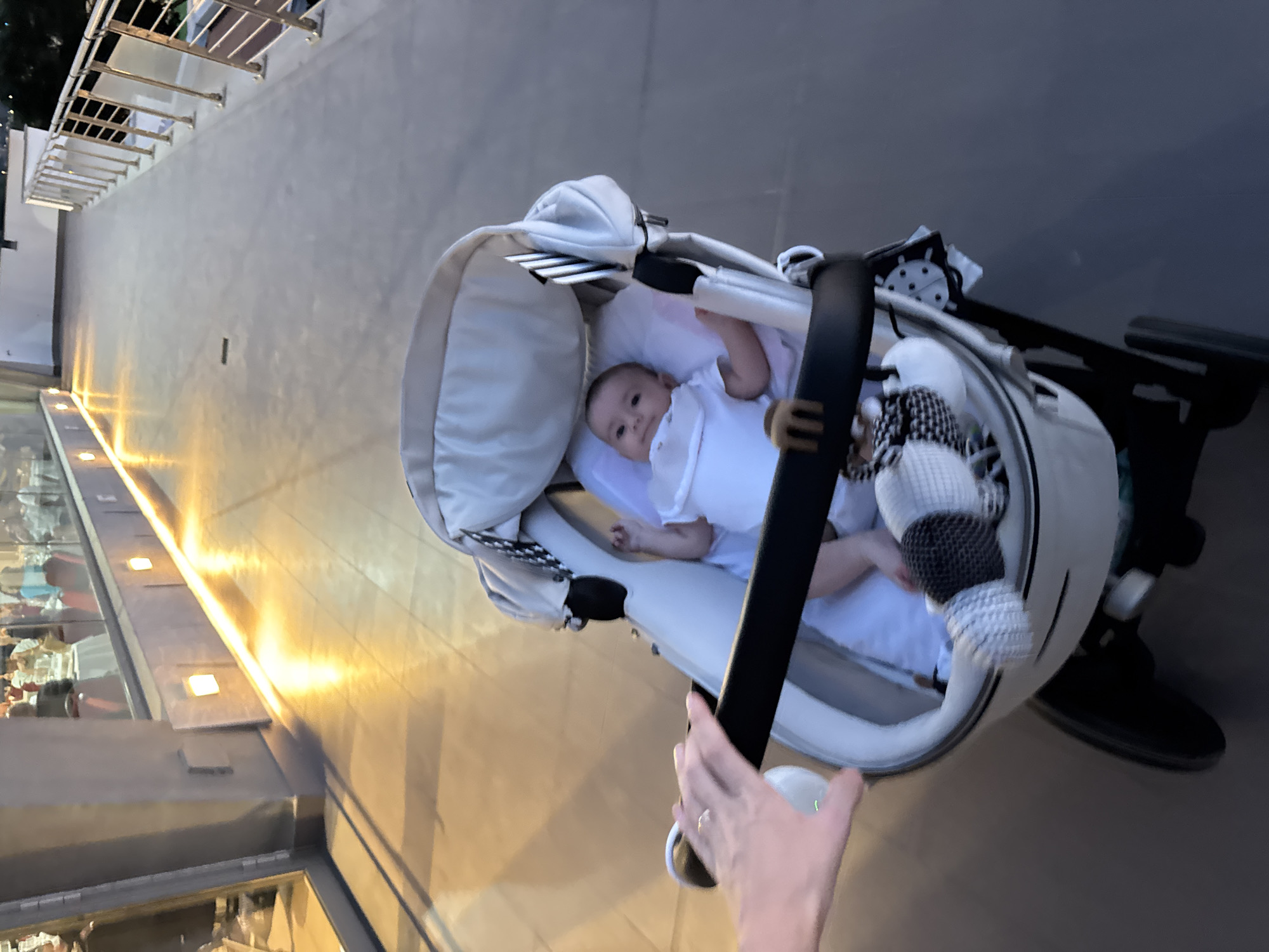 A baby is in a stroller on a sidewalk in front of a building with glass windows and outdoor lighting. The baby is wearing a white outfit and bib. A hand is visible on the stroller handle. The sidewalk is paved with rectangular tiles. The building's exterior is made of concrete with recessed lighting. A metal railing lines the edge of the sidewalk. The background is blurry, but appears to be an outdoor setting at dusk. The location is identified as Isola delle Femmine, Italy.