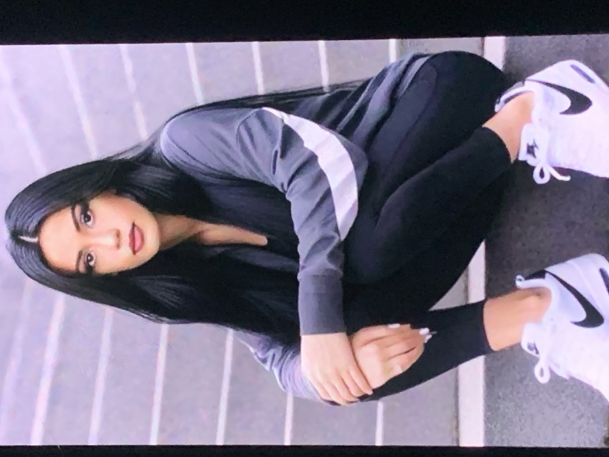 A young woman with long, dark hair is sitting on concrete steps outdoors. She is wearing a grey jacket with white stripes on the sleeves, black leggings, and white Nike sneakers. Her hands are crossed in front of her. The background consists of layered concrete steps. The scene appears to be a public space in Sevastopol, Ukraine.