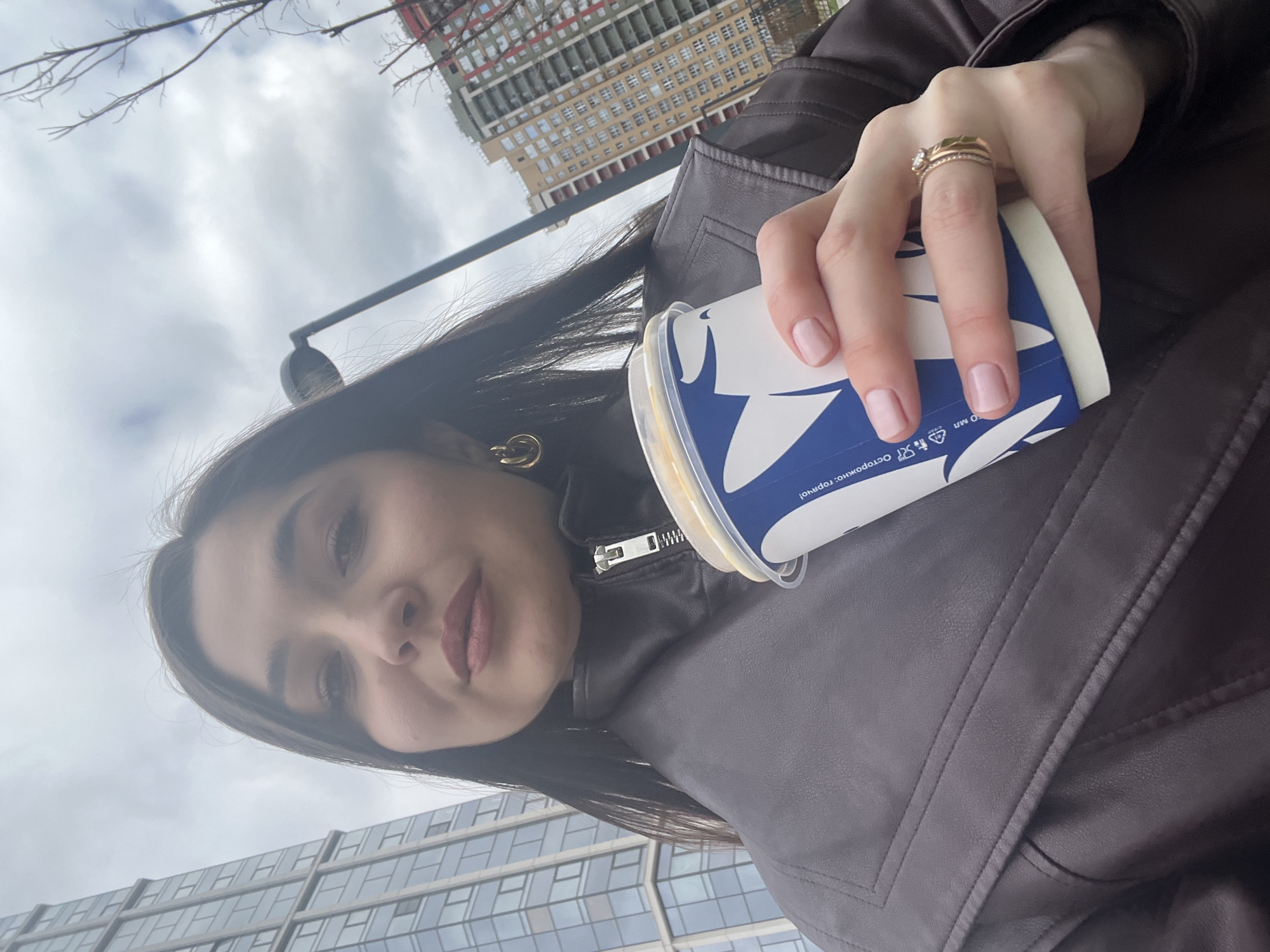 A tilted outdoor selfie features a young woman holding a beverage cup on an overcast day in Khimki, Russia. Her face, seen from a low angle, occupies the left and center of the frame, showing dark hair, dark eyebrows, and dark lipstick, with a neutral to slightly smiling expression. She is wearing a dark brown or black leather-look jacket with a visible zipper and stitched details.
Her left hand, adorned with two gold-toned rings on her ring finger (one with a prominent stone, another a simple band), holds a white cup with a blue and white design. A clear plastic lid tops the cup, revealing a yellowish liquid inside. Visible blue text on the cup reads "ОСТОРОЖНО: ГОРЯЧО!" (CAUTION: HOT!), accompanied by recycling symbols and a "do not litter" icon. She has light pink nail polish and a gold-colored, circular earring in her left ear.
The background shows an urban environment under a cloudy sky. To the upper right, a tall, light-colored building, possibly under construction, features many windows. Another modern building with large glass windows is visible below her face. Bare tree branches are seen in the upper left, and a dark pole, potentially a street light, extends from the upper right, partially obscured by her hair.