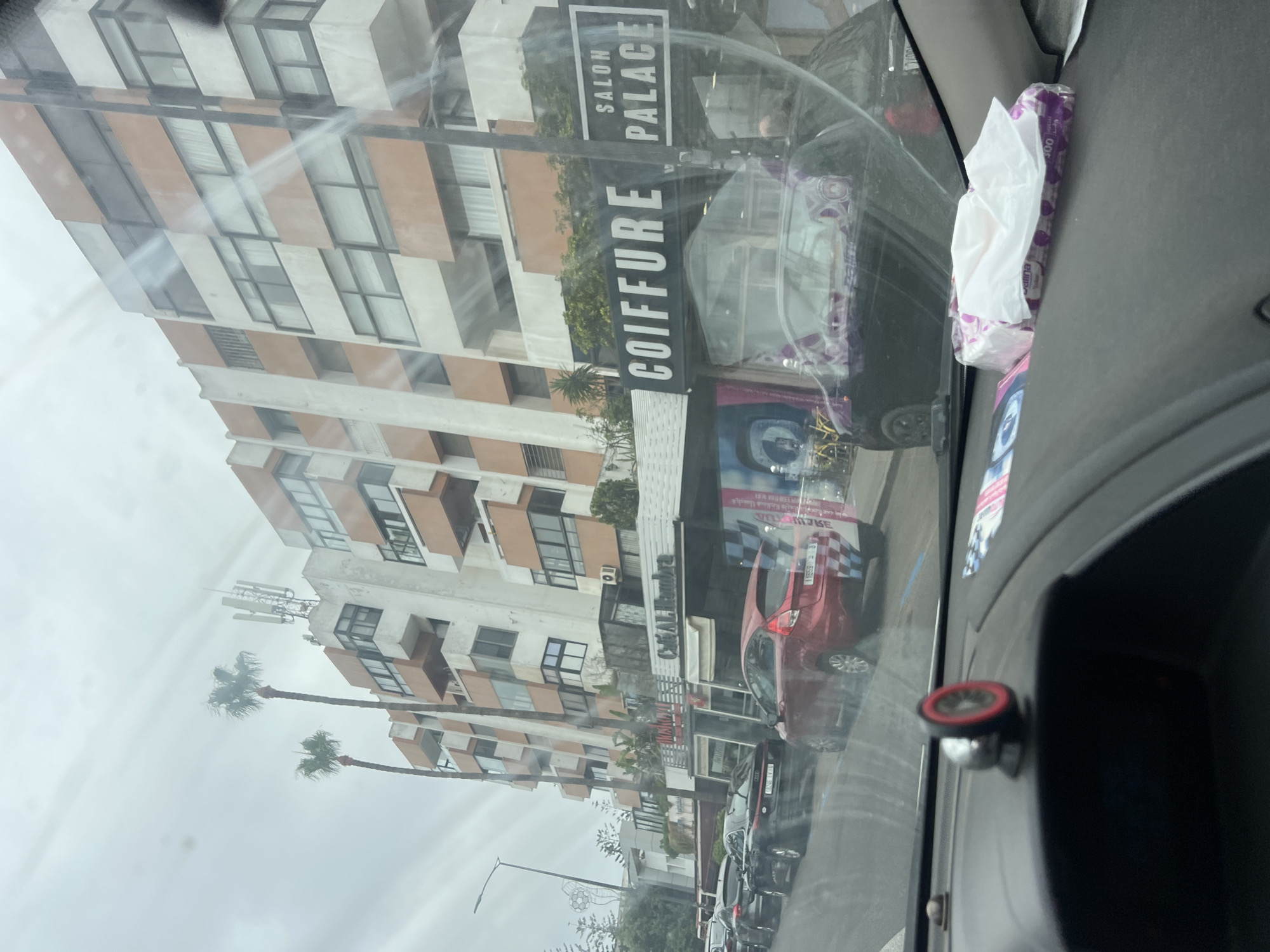 The image presents a street-level view from the interior of a vehicle on Boulevard Ghandi in Casablanca, Morocco, captured on an overcast day. The foreground shows the car's dashboard, featuring a red and black magnetic phone mount and a white and purple pack of Selina tissues positioned against the windshield. Reflections on the windshield obscure parts of the view but reveal elements of the vehicle's interior.
The street is lined with parked vehicles and multi-story buildings with a distinctive architectural style characterized by white facades interspersed with horizontal and vertical orange-brown panels and balconies. Two prominent palm trees rise above the buildings, and a tall cellular antenna structure is visible on the rooftop of one building.
On the ground floor, several commercial establishments are present. A large sign for "COIFFURE SALON PALACE" is clearly visible, indicating a hair salon. Adjacent to it, another sign reads "CAFÉ ALHAMBRA." Further down the street, another business sign is partially visible, potentially reading "NOSTALGI..." Several cars are parked along the curb, including a black Audi sedan with the Moroccan license plate "1246 | Y | 18" and a red Ford Fiesta hatchback displaying the Moroccan license plate "11559 | ب | 61." No individuals are clearly discernible in the scene.