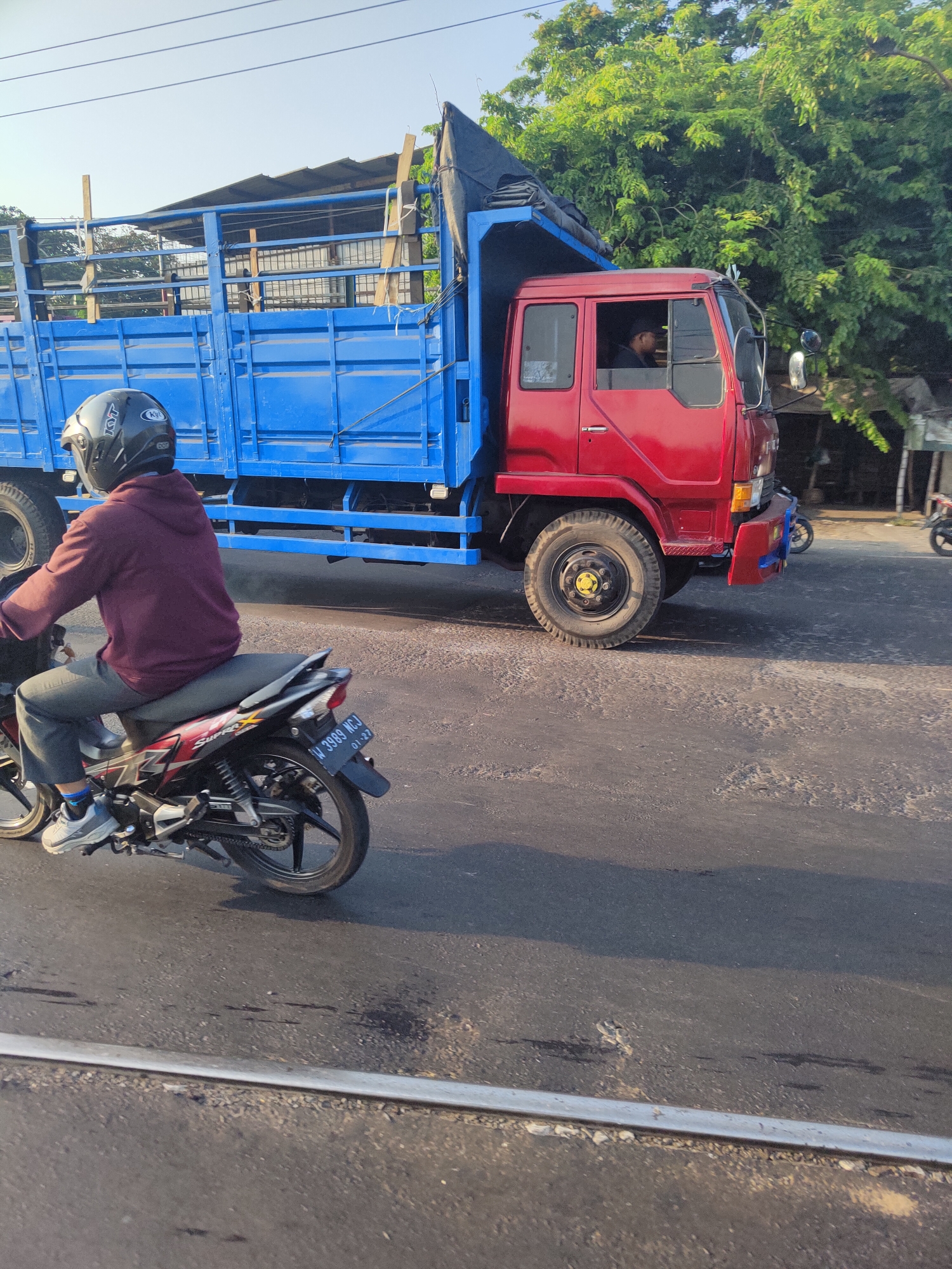 A motorcycle carrying a person in a maroon hoodie and a helmet rides on a paved road. The motorcycle has a license plate with the characters "W 3989 WCJ" and the number "01-27". Next to the motorcycle, a red truck with a blue cargo bed is driving on the same road. A person is visible in the driver's seat of the truck. Behind the truck, lush green trees are seen. The sky is clear and blue. The scene appears to be an outdoor environment, possibly in Latek, Indonesia, given the style of the vehicles and the foliage.