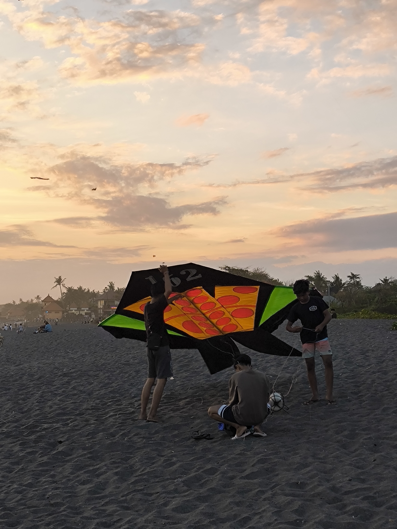 A group of young men are on a dark sand beach in Mengwi, Indonesia, preparing to fly a large, colorful kite. The sky above is filled with soft, pastel-colored clouds, indicative of a sunset. Two of the men are standing, one holding the kite's string and the other assisting with the kite. A third man is crouched down, tending to the kite's string and a ball. In the background, palm trees and buildings are visible, suggesting a coastal resort area. Other beachgoers can be seen in the distance.
