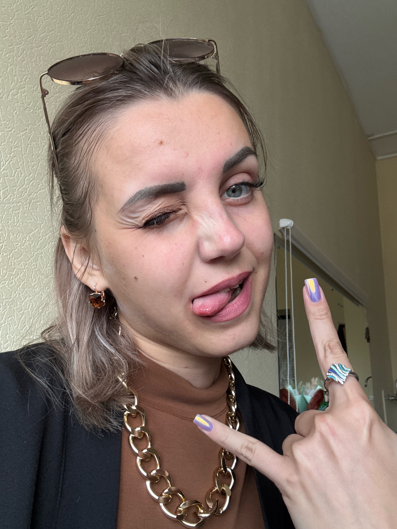 A young woman is pictured in a medium shot, indoors, likely in an apartment in Yuzhno-Sakhalinsk, Russia. She is looking at the camera with a playful expression, winking her left eye and sticking out her tongue. She has blonde hair styled back with sunglasses perched on top of her head. She is wearing a black blazer over a brown turtleneck, a thick gold chain necklace, and large brown earrings. Her fingernails are painted with a purple and yellow design. She is wearing a silver ring with blue and white wave-like embellishments on her right index finger. The background shows a textured wall and a glimpse of a mirror.