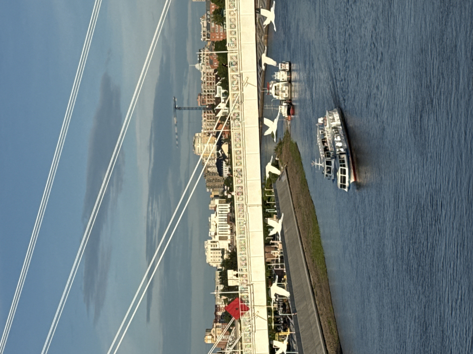 A cable-stayed bridge with white cables spans a river in Tyumen, Russia. The bridge has a white deck with decorative railings. There are large, white bird sculptures mounted on the side of the bridge's support structure. Several boats are visible on the river, including a passenger boat in the foreground and a group of smaller vessels docked further upriver. In the background, a city skyline with various buildings is visible, along with a construction crane. The sky is partly cloudy, with a bright blue expanse. A red square-shaped object is visible on the bridge. The riverbanks are lined with concrete structures and some greenery.