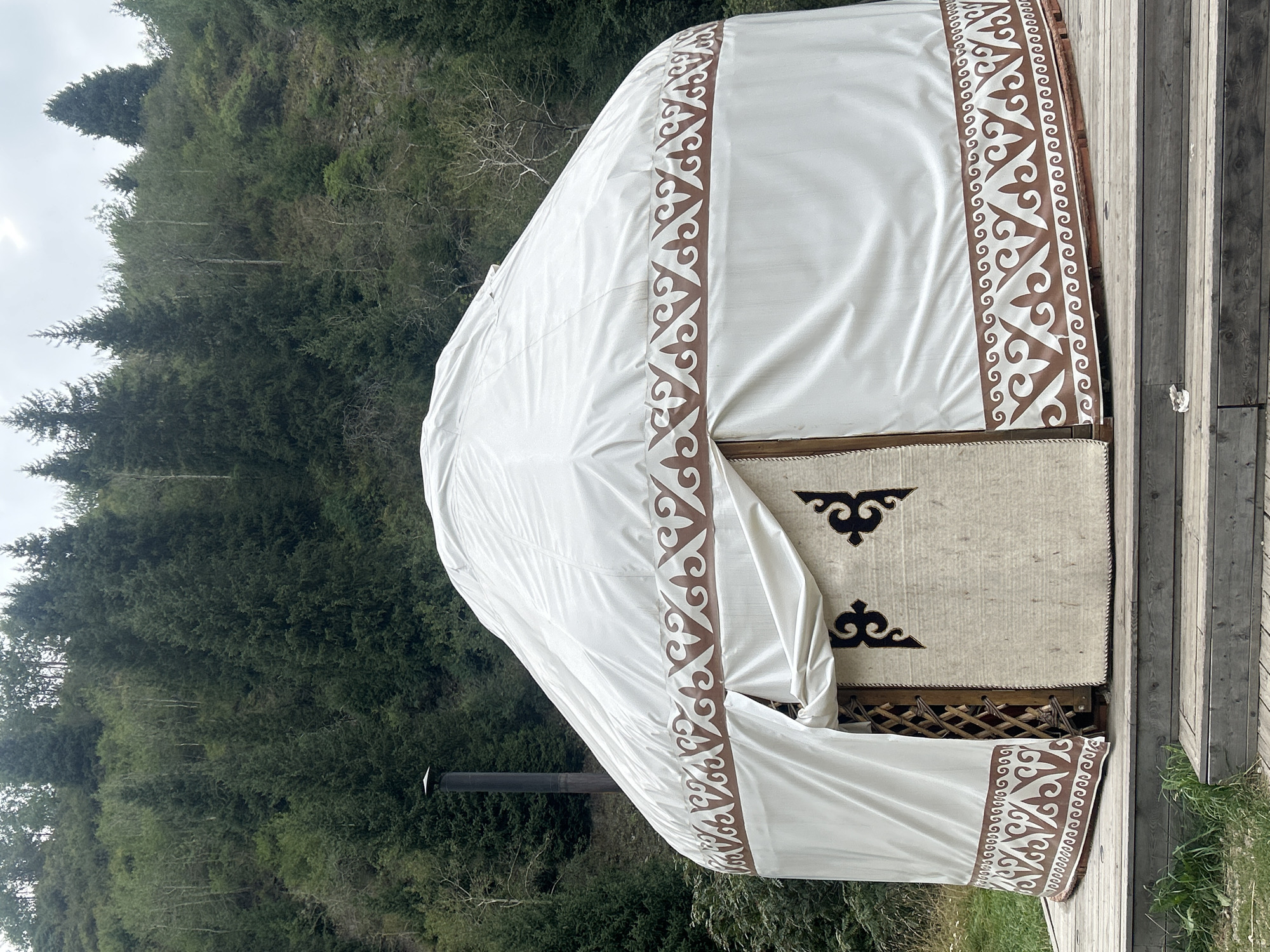 A yurt with a white exterior and brown decorative trim sits on a wooden platform. A chimney pipe extends from the left side of the yurt. The yurt is situated in a mountainous area with a dense forest covering the hillside behind it. The sky is overcast. This scene is located in Almaty, Kazakhstan.