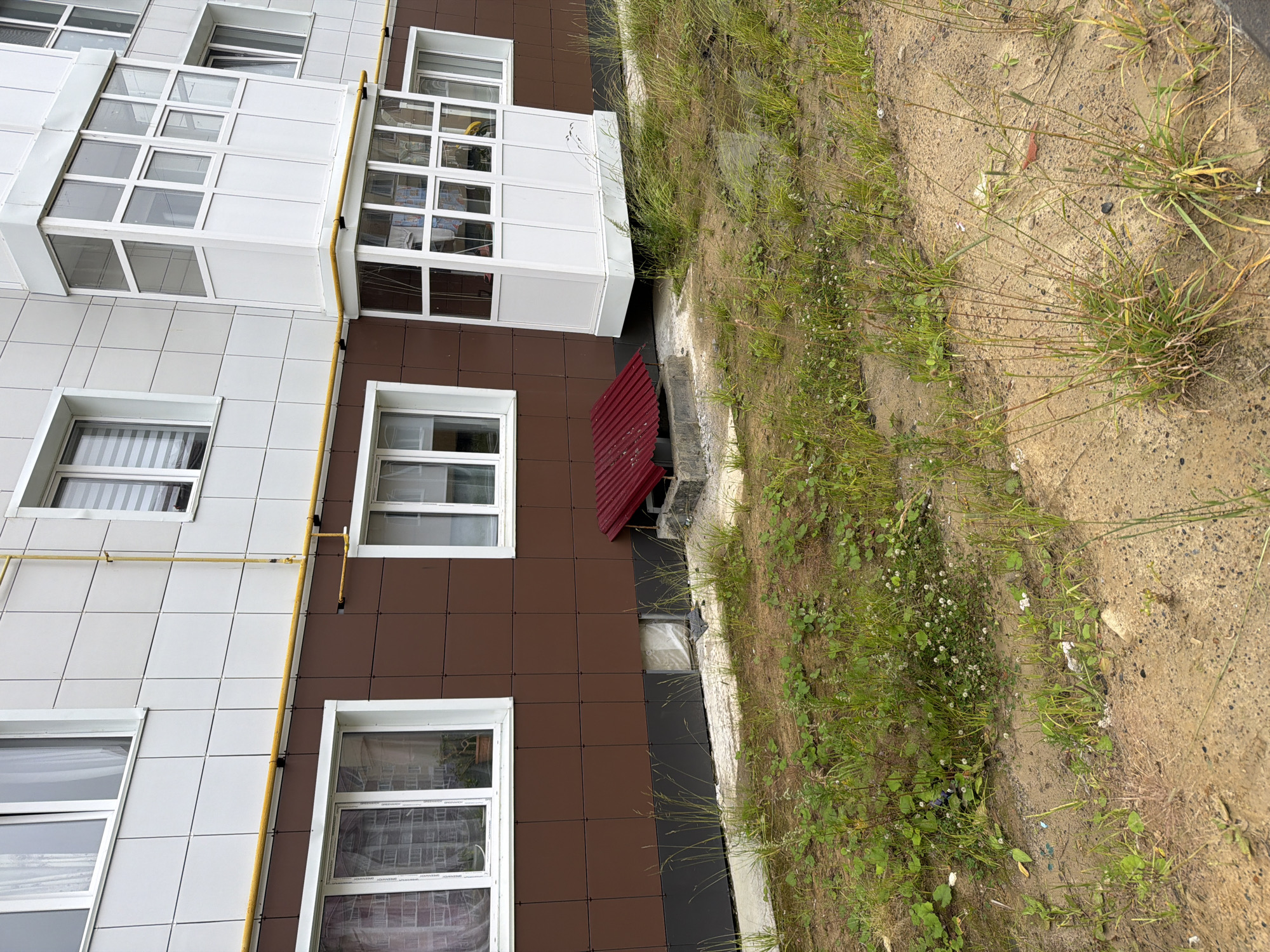The image depicts the exterior of a multi-story building in Okha, Russia. The building features white tiled upper levels and brown tiled lower levels. A yellow gas pipe runs horizontally along the building. There are several windows visible, some with curtains. A white enclosed balcony or sunroom is attached to the building. In front of the building, on the ground level, is a small, red, corrugated metal structure, possibly a ventilation cover or access point. The area in front of the building is partially paved with concrete and partially covered with dry soil and overgrown grass, indicating an unkempt outdoor space. There are no people visible in the image.