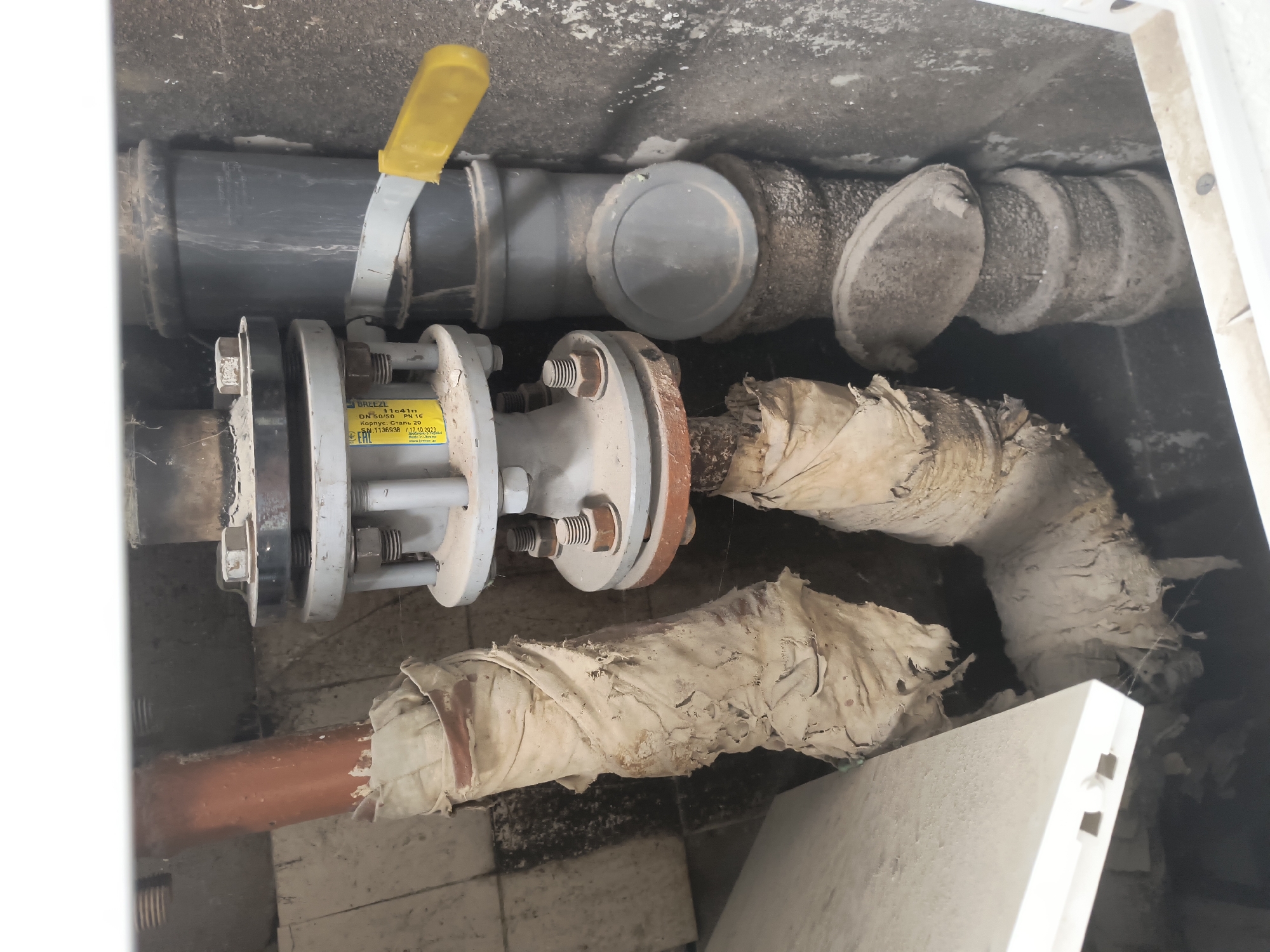 The image depicts a close-up view of plumbing infrastructure, likely within a utility access point or basement. The central focus is a gray industrial valve assembly with a yellow lever handle, connecting two sections of pipe. To the left, a reddish-brown pipe, partially wrapped in deteriorating, light-colored insulation material, is visible. To the right, a larger, dark gray pipe is also present, with additional pipe sections and fittings below it. The surrounding environment appears to be a rough-hewn, possibly concrete or masonry, enclosure with a dusty and somewhat grimy surface. A white, flat object, possibly a cover or part of the enclosure, is visible on the left side of the frame. The overall impression is of a functional, but somewhat aged and weathered, piece of industrial equipment. There are no people or specific identifiable landmarks related to Rivne, Ukraine, in this image.