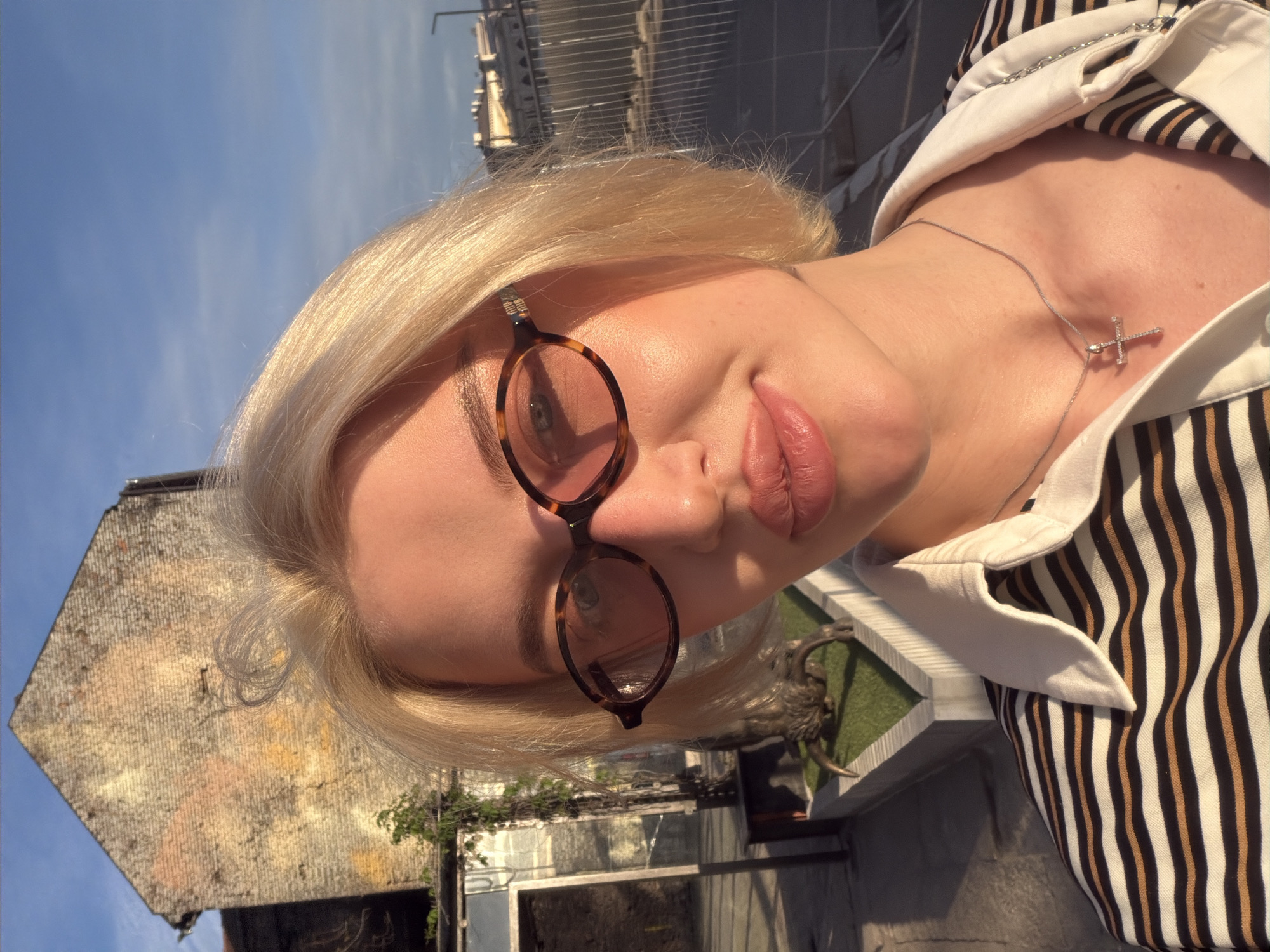 A blonde woman with sunglasses and a striped shirt poses in a close-up shot. Behind her is a building with a weathered roof and a building behind a railing. The buildings are located in Belgrade, Serbia. She wears a delicate silver necklace with a cross.