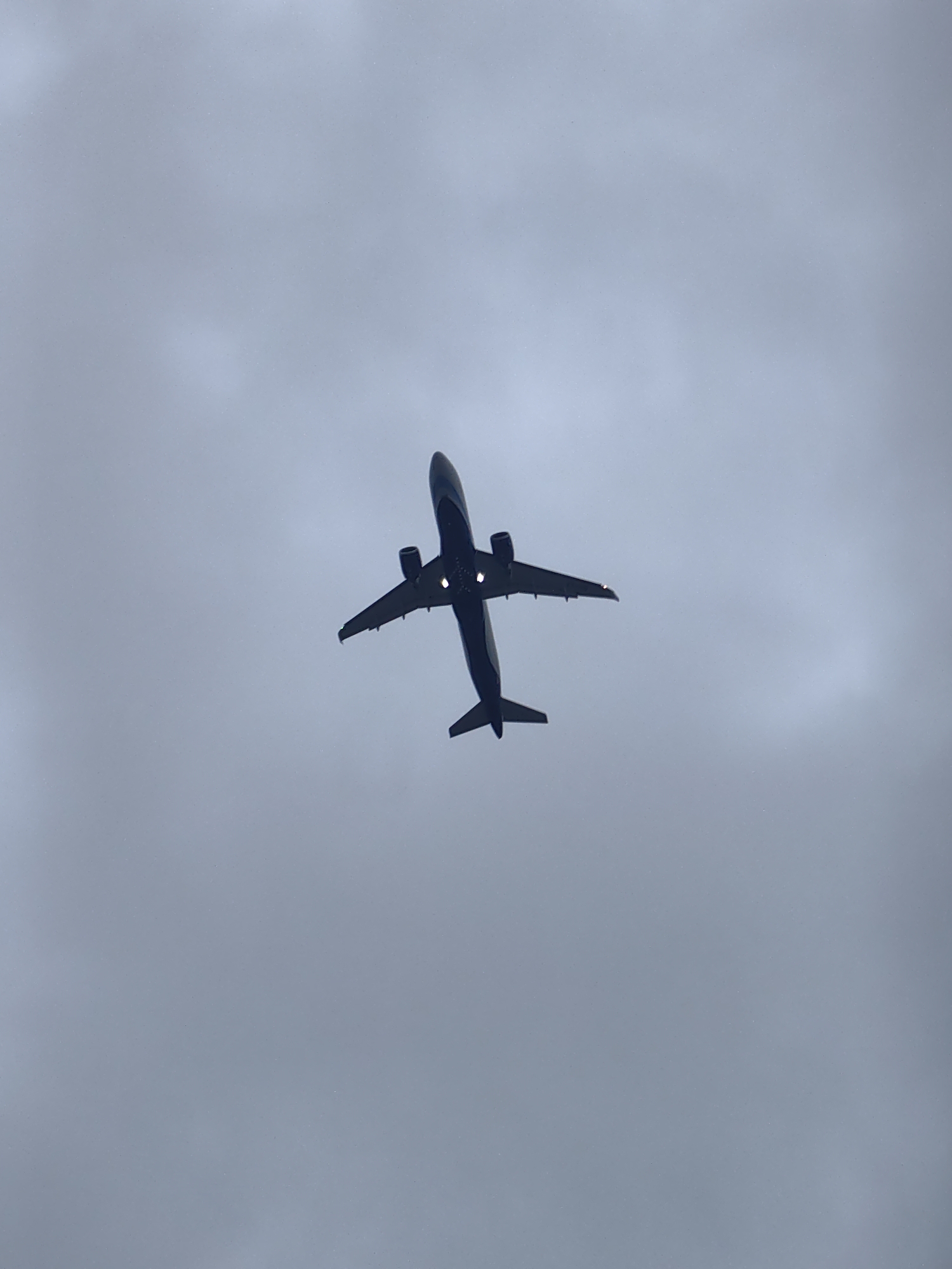 A wide shot shows an airplane viewed from directly below, ascending against a dull, overcast sky. The aircraft is dark blue with visible white lights on its underside. Its wings are extended and angled upwards, and the tail is clearly visible. The sky is a uniform light gray with subtle cloud formations, suggesting a cloudy or foggy day. The overall impression is one of an aircraft in flight over an unseen area.