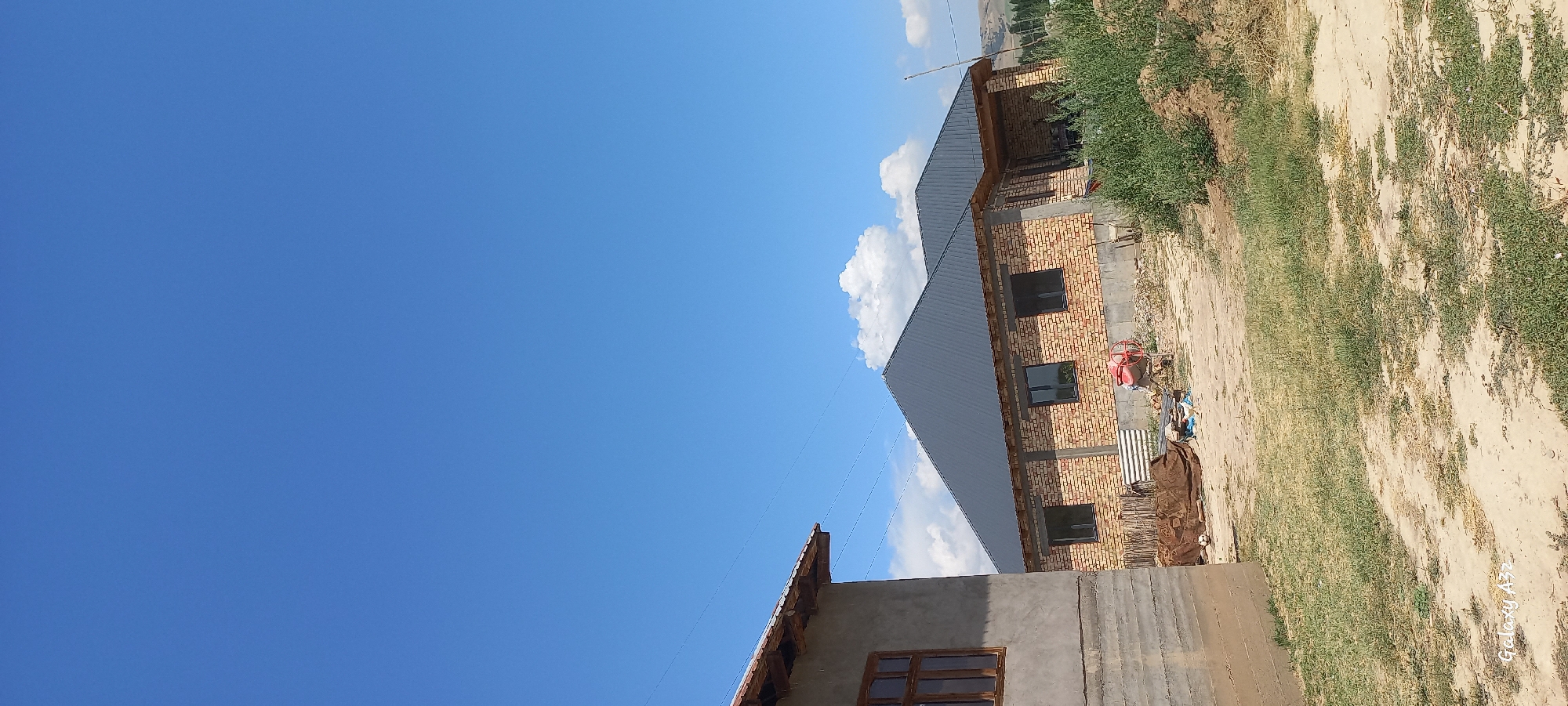 In the city of Joon-Kunggöy, Kyrgyzstan, two unfinished brick houses stand under a clear blue sky. The house on the left is made of concrete with a wooden window frame. The house on the right, constructed with yellow bricks, features a dark gray metal roof and dark-framed windows. Construction materials, including a concrete mixer, are scattered around the site. Dry grass and dirt cover the ground, with a line of green vegetation visible in the foreground. The background reveals rolling hills under a vast expanse of blue sky dotted with white clouds.