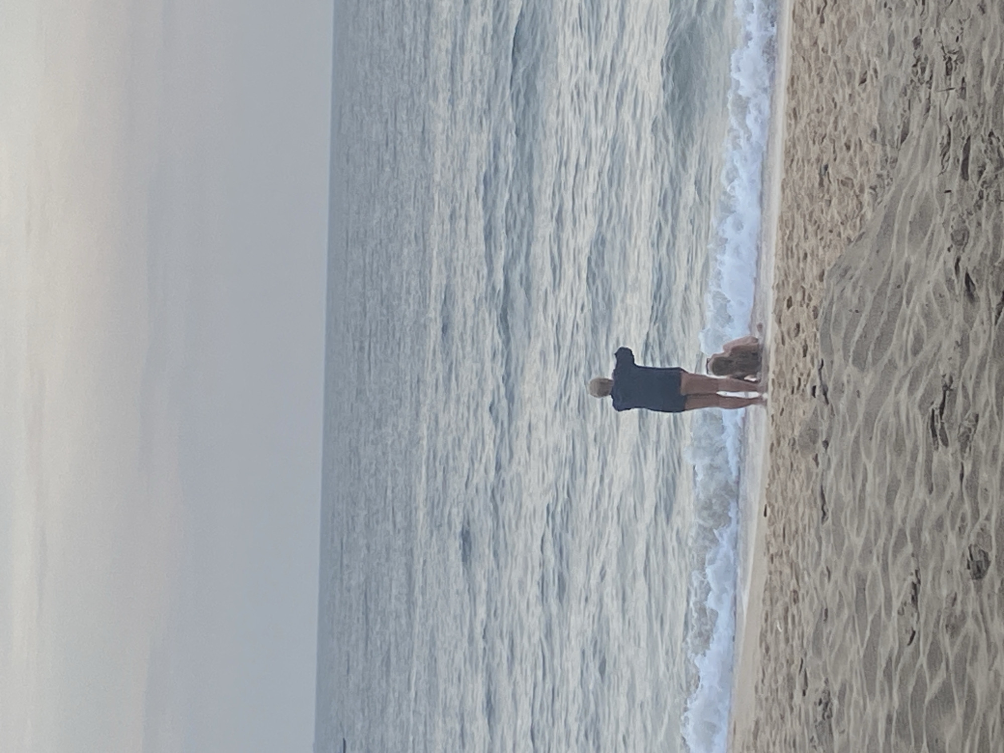 An overhead drone shot captures two individuals on the beach in Mũi Né, Vietnam. A person stands near the water's edge, facing the ocean with their arms outstretched as if performing a gesture. Another person is seated or lying down in the shallow water. The beach sand is textured with ripples. The ocean is calm with gentle waves breaking at the shore, and the sky is overcast.
