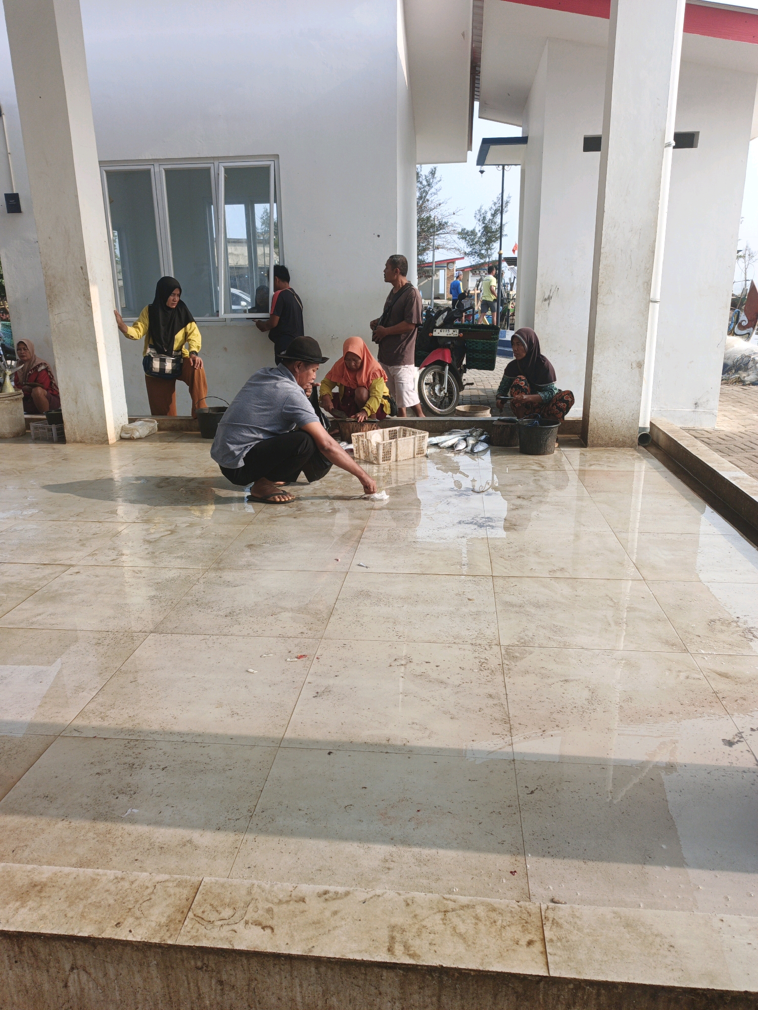 In an outdoor covered area with tiled flooring, several individuals appear to be engaged in the handling of fish. The scene suggests a marketplace or processing area, likely near a body of water given the presence of fish and the overall atmosphere. The flooring is wet and reflects the overhead structures and sunlight, indicating a humid or recently cleaned environment.
In the foreground, a man wearing a dark hat and a gray t-shirt is squatting and reaching towards the floor, seemingly examining or picking up something. To his right, a woman in an orange headscarf and yellow top is also squatting, facing a white crate filled with fish.
Further back, a woman in a yellow headscarf and dark clothing stands near a window, looking towards the activity. To her left, another woman in a red garment is also squatting, next to a white crate.
In the mid-ground, a red motorcycle is parked. Behind the motorcycle, more individuals are present, including a man in a dark shirt and shorts. Another woman in a dark headscarf is seen squatting near the motorcycle, and appears to be working with a chopping board and fish.
The background shows more of the covered structure, trees, and a glimpse of what could be a body of water or beach in the distance. The lighting suggests it is daytime, possibly late morning or afternoon. The overall impression is a busy, working environment where fish are being prepared or sold.