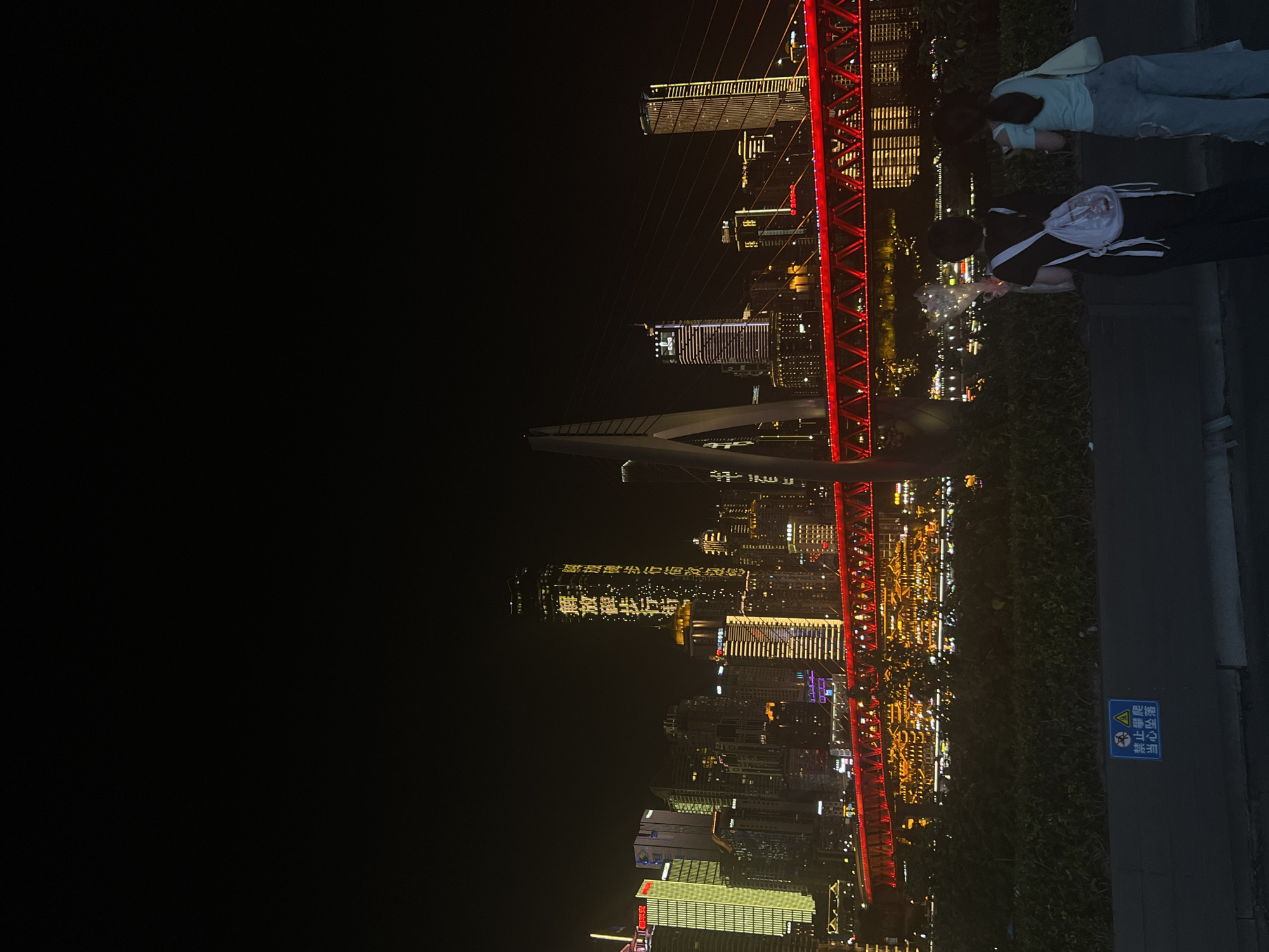 The image depicts a nighttime cityscape in Jiangbeicheng Street, China, dominated by illuminated skyscrapers and a prominent red bridge. Two individuals, a female with dark hair wearing a black top and backpack, and another female with dark hair in a light blue top and jeans, stand facing away from the camera, looking towards the illuminated cityscape. The female on the left holds a bunch of illuminated objects, possibly lights or glowing decorations. The bridge, spanning across a body of water, is a vibrant red structure with intricate supports and lighting. In the foreground, there is a dark green vegetated area and a gray wall with a blue sign displaying Chinese characters and symbols. The sky is dark and clear, characteristic of a night scene. The buildings in the background are a mix of modern high-rises, some with illuminated vertical text, and other structures with extensive lighting. The overall atmosphere is one of a bustling urban environment at night, with a focus on the architectural illumination and the presence of pedestrians.