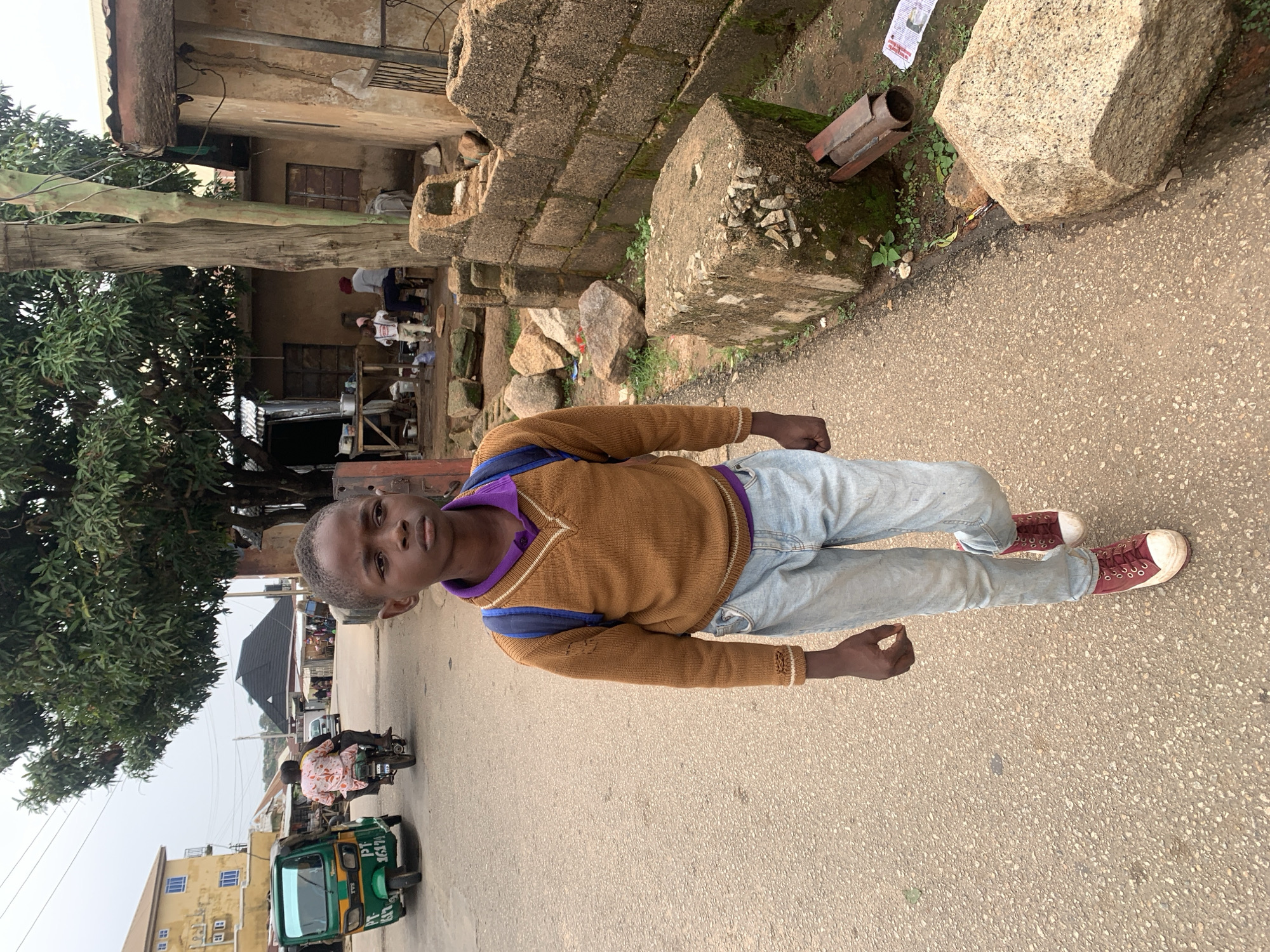 A young boy, dressed in a brown knitted sweater with a purple collar and light blue jeans, stands on a paved road in what appears to be Jos, Nigeria. He is facing the camera with a neutral expression. The boy is wearing maroon high-top sneakers. Behind him, a lush green tree is visible, and further in the background, there are buildings and people on motorcycles. The scene suggests a daytime setting, possibly in the late afternoon, with overcast skies. The environment seems to be a mix of urban and somewhat rural elements, with some stone structures and vegetation. A green auto-rickshaw with "PT 1617" visible on its side is in the foreground, alongside another motorcycle with a passenger. The overall impression is of a casual street scene in a Nigerian town.