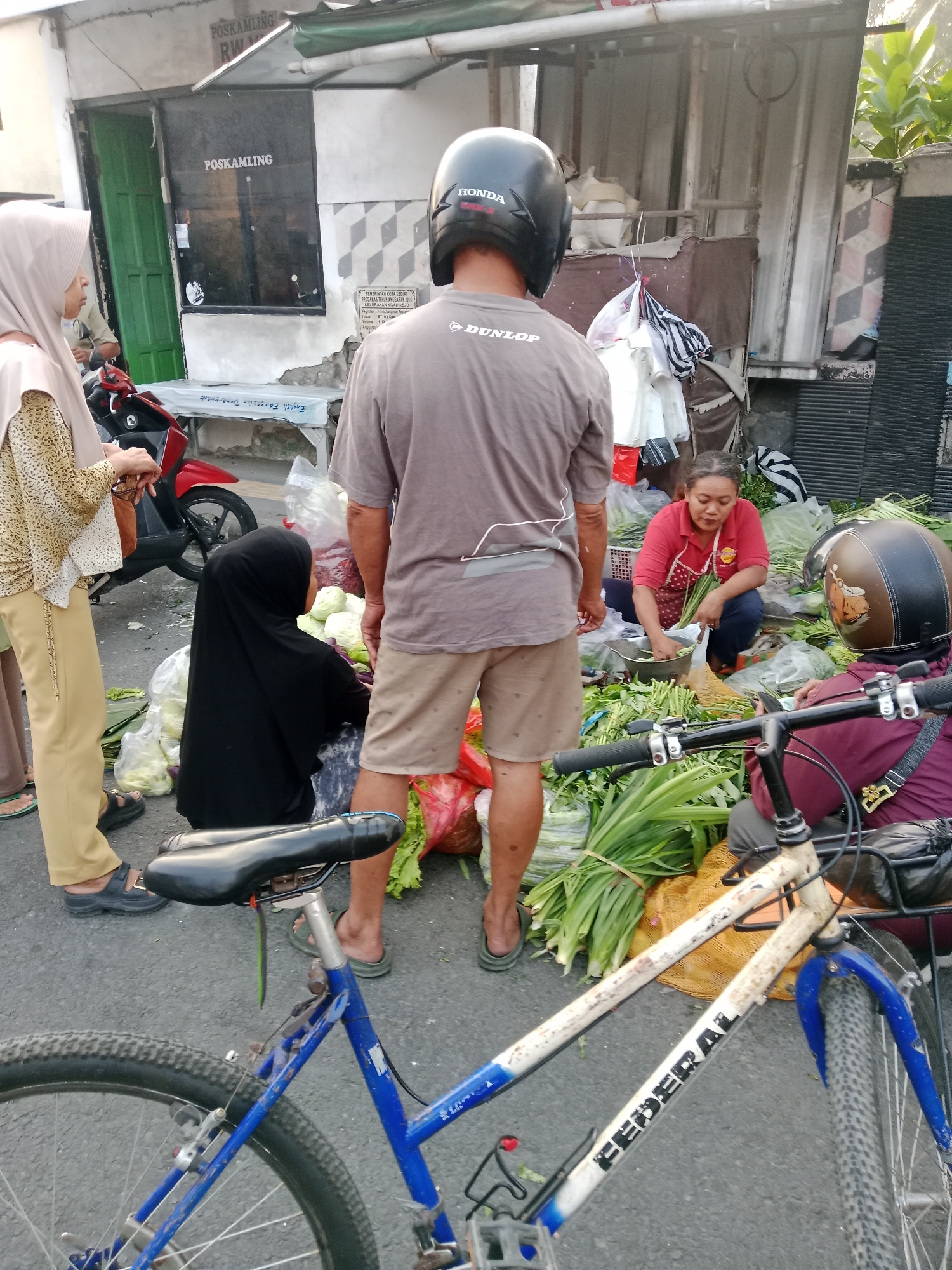 The image depicts a street scene in Kediri City, Indonesia, with several people and objects. In the foreground, a blue bicycle is prominently displayed, with its front wheel and frame visible. A person wearing a burgundy shirt and a helmet is seated on a motorcycle behind the bicycle. To the left, a woman in a beige hijab and patterned blouse is standing near a red motorcycle. Behind her, a young person in a black hijab is seated among piles of vegetables. Further back, a woman in a red shirt and apron is busy preparing vegetables, with a pot and more produce around her. Several other individuals are present in the background. The setting appears to be a marketplace or roadside vendor area, with various items for sale, including fresh produce. The overall environment suggests a bustling, outdoor commercial activity. A building with the sign "POSKAMPLING" is visible in the background, indicating a local community post or security point.