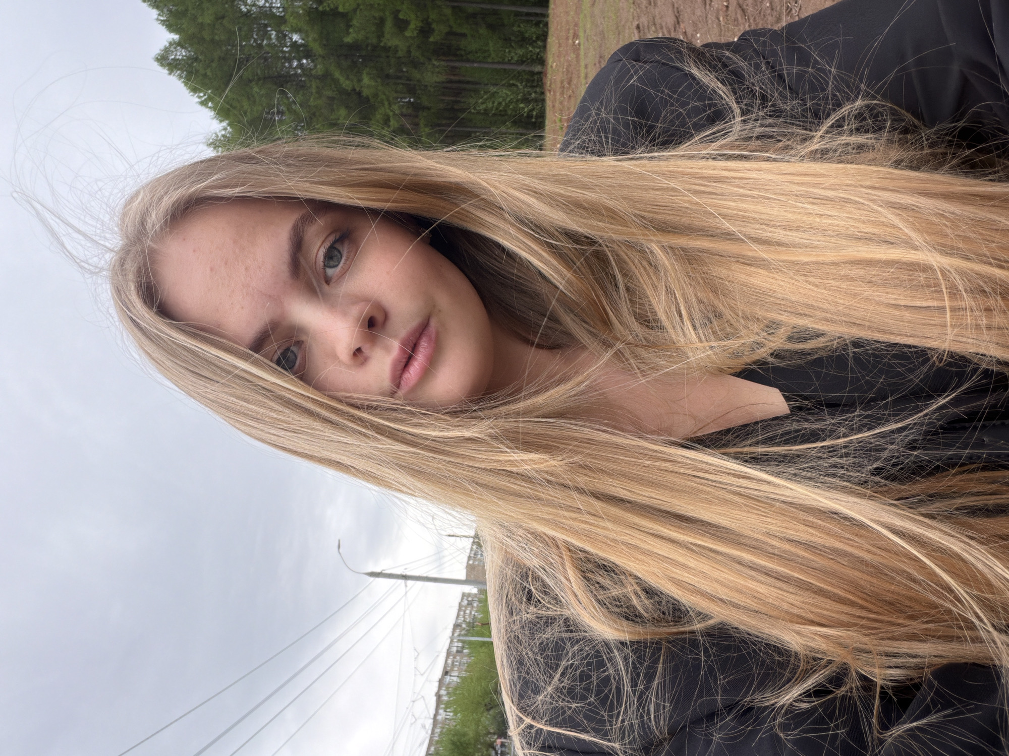 A selfie image shows a young woman with long, blonde hair framing her face. She has fair skin and is wearing a black shirt. The background includes a cloudy sky, trees, and a bridge structure with cables, appearing to be a suspension bridge. The location is Izhevsk, Russia.