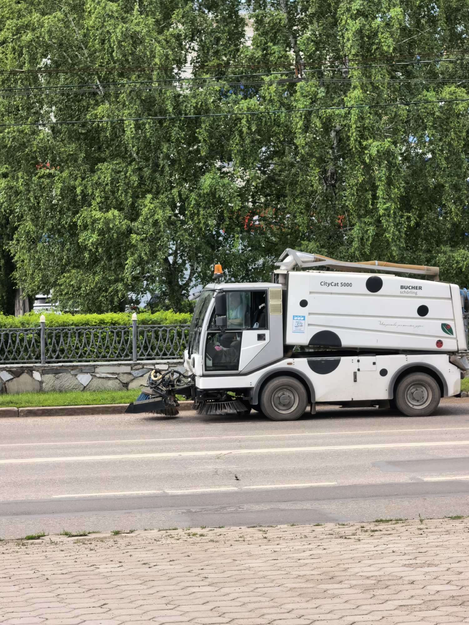 The image shows a white Bucher CityCat 5000 street sweeper truck operating on a paved road. The truck has a prominent cylindrical debris container and side brushes. A person is visible in the driver's cabin. The truck is positioned on the right side of the road, moving from left to right. In the background, there is a green metal fence, a hedge, and a dense canopy of green trees with some power lines running through them. The road surface is asphalt with white lane markings. The foreground shows a sidewalk paved with interlocking concrete blocks, with some sparse vegetation growing between them. The scene appears to be in an urban environment, likely a street in Sterlitamak, Russia, given the context of the query. The lighting suggests it is daytime, possibly overcast or late afternoon.