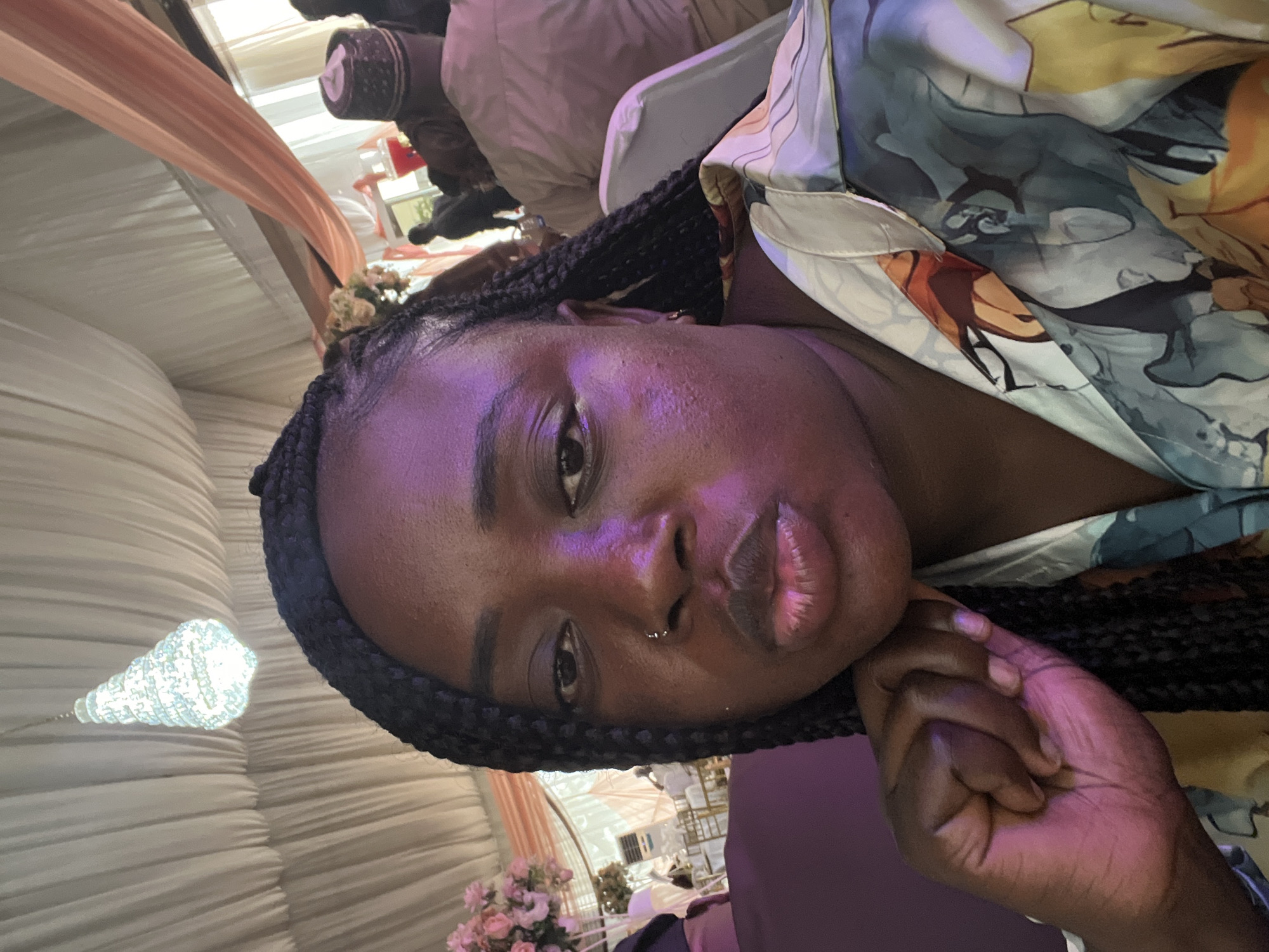 A close-up, slightly rotated photograph shows a dark-skinned woman with braided hair, wearing a patterned shirt, at what appears to be a wedding reception. The woman is resting her cheek on her hand.
In the background are white draped walls, floral decorations, a chandelier-type light fixture, and glimpses of seated people.
Given the presence of traditional wedding elements, and the likelihood of such events occurring in Abuja, Nigeria, it is likely this photo was taken in Abuja. There is no additional data to support this likelihood.