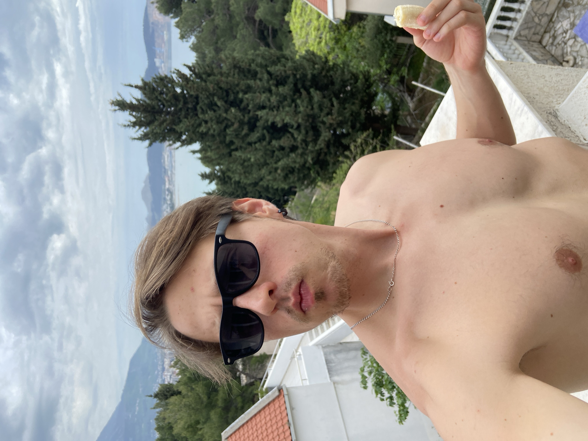 A shirtless man wearing sunglasses is holding a partially eaten banana. He has light brown hair and is wearing a silver chain necklace. The background includes greenery, buildings, and a distant view of mountains and the sea. The location is Šušanj, Montenegro.
