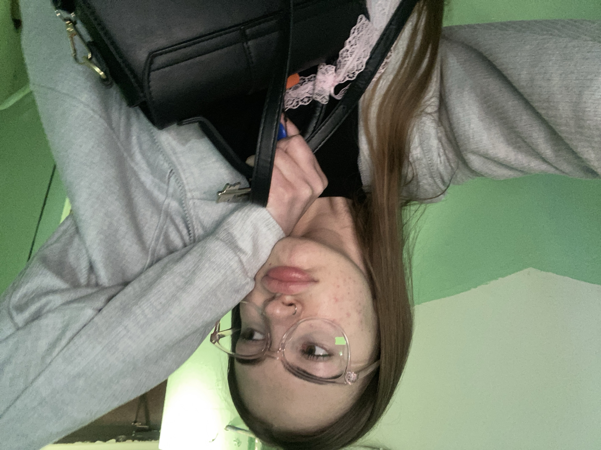 A head-and-shoulders selfie depicts a person with long, straight brown hair, wearing large, translucent, light-colored framed eyeglasses and a silver hoop nose piercing on the left nostril. The subject displays a pronounced pout, with their head resting on their bent left arm, which is clad in a light grey hooded sweatshirt. The facial skin exhibits multiple blemishes. The right hand, also covered by the grey sweatshirt sleeve, holds the handle of a black structured handbag featuring a flap closure and gold-toned hardware. Visible within or attached to the bag are a blue pen, an orange object, and a piece of light pink lace trim. The background consists of an interior wall painted with a diagonal division: the upper section is a light, muted green, and the lower section is a darker, more saturated green. Indistinct dark elements are present in the upper right background. The location is Unknown, Unknown.