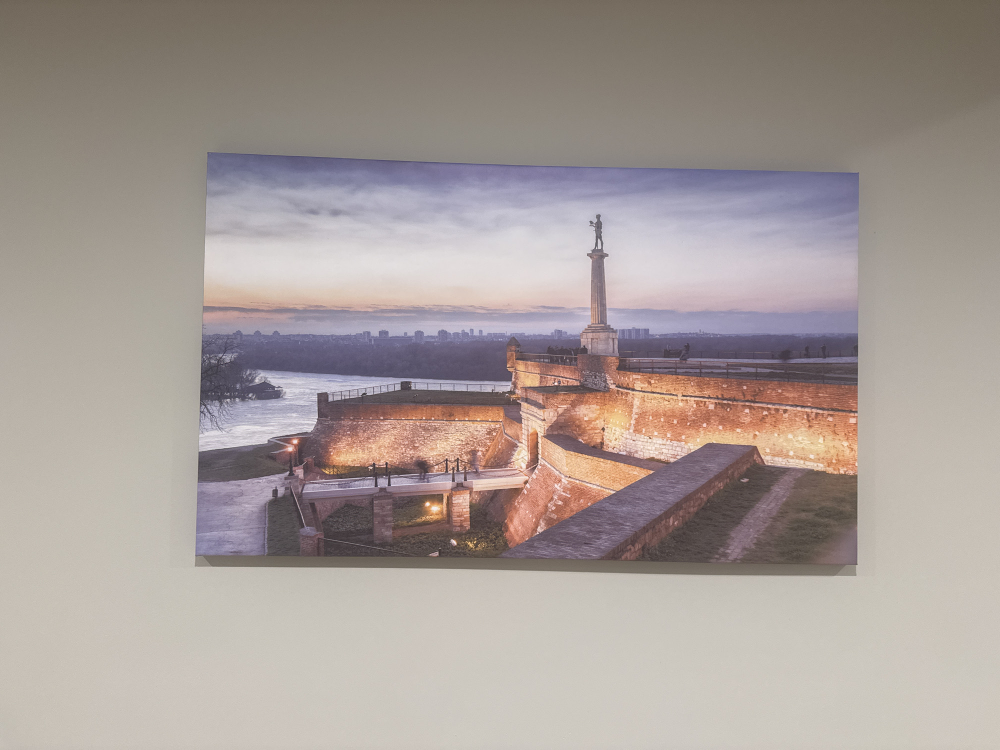 The image depicts a large canvas print of the Pobednik monument and Belgrade Fortress at sunset. The monument, a prominent statue on a stone column, stands tall against a dusky sky with hues of purple, pink, and orange. Below the monument, the brick walls of the fortress are illuminated, casting a warm glow. A bridge with a walkway leads across a grassy area towards the fortress entrance. In the distance, the city skyline of Belgrade is visible, with a river flowing in the foreground. The canvas is mounted on a light-colored wall, with the lighting in the room creating a subtle shadow on the wall behind the print.