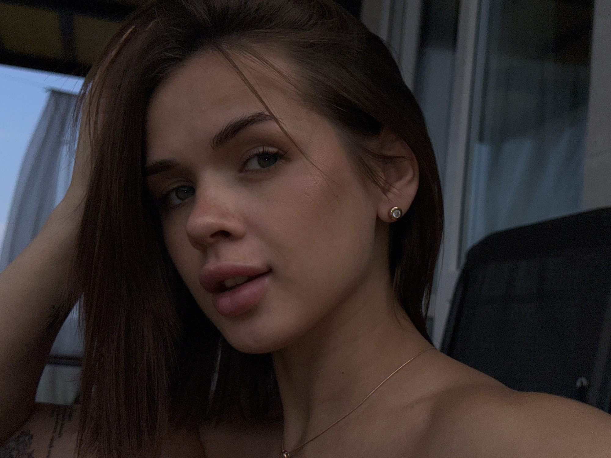 A close-up, shoulder-up portrait of a young woman with shoulder-length brown hair. She is wearing a small, gold stud earring with a crystal. Her head is tilted slightly, and her left hand is raised to her hair. She has light skin and dark eyebrows. Her lips are slightly parted, and she is wearing subtle makeup. A thin gold necklace is visible around her neck. The background is out of focus and appears to be an outdoor setting with some structures suggesting a balcony or patio area, possibly with a view of a blue sky. There is a dark object behind her on the right side of the frame, which could be furniture or part of the building structure. No specific landmarks are visible to definitively place the image in Lobanovo, Russia.