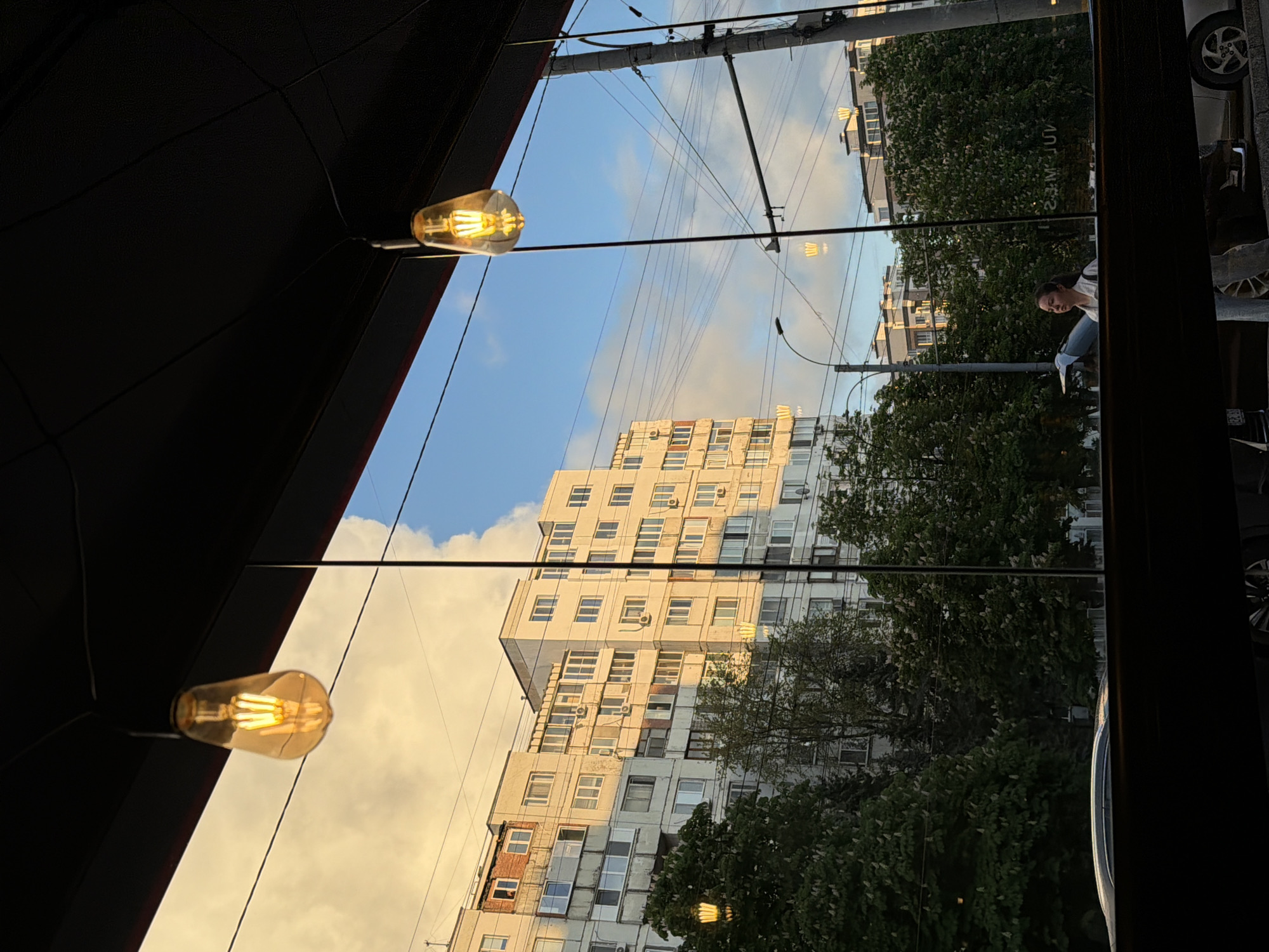 The image shows an outdoor scene viewed through a window. Two light bulbs hang from the dark ceiling of an indoor space. Beyond the window, a tall, white apartment building stands. Green trees are situated between the building and the window. Power lines stretch across the sky, which is blue with white clouds. Reflected in the window is a woman wearing a white shirt and blue jeans. A car is also visible in the window reflection. The location appears to be a city in an unknown location.