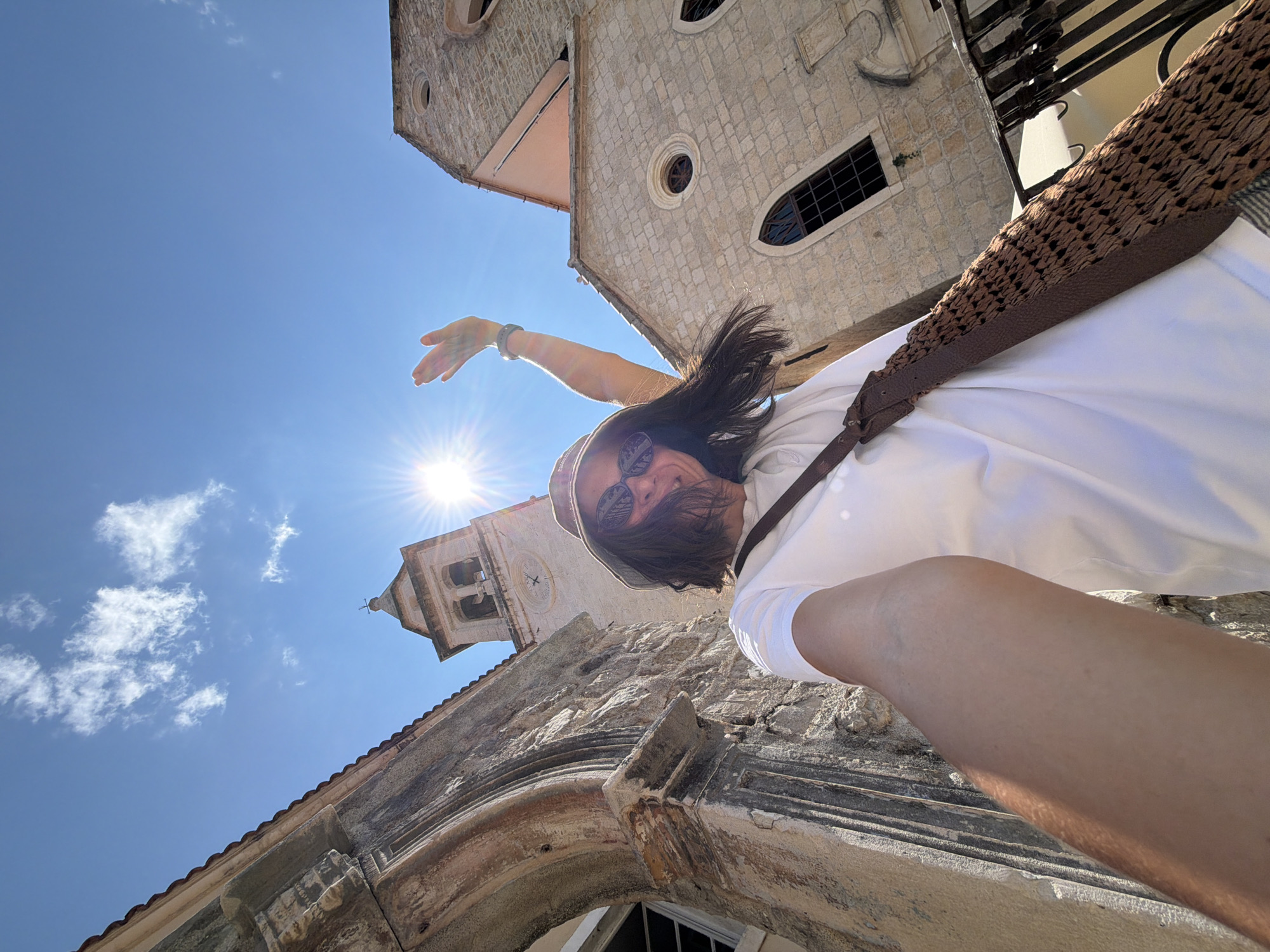 A person is taking a selfie in front of a stone building with a bell tower and a clock. The person is wearing a white t-shirt, sunglasses, and a hat. They are holding a brown woven bag across their chest and waving with their right hand. The sun is shining brightly, creating a lens flare. The architecture suggests a European city, possibly a historic old town area. Based on visual cues consistent with the architecture, bell tower style, and building materials, this scene is likely located in the Old Town of Budva, Montenegro.