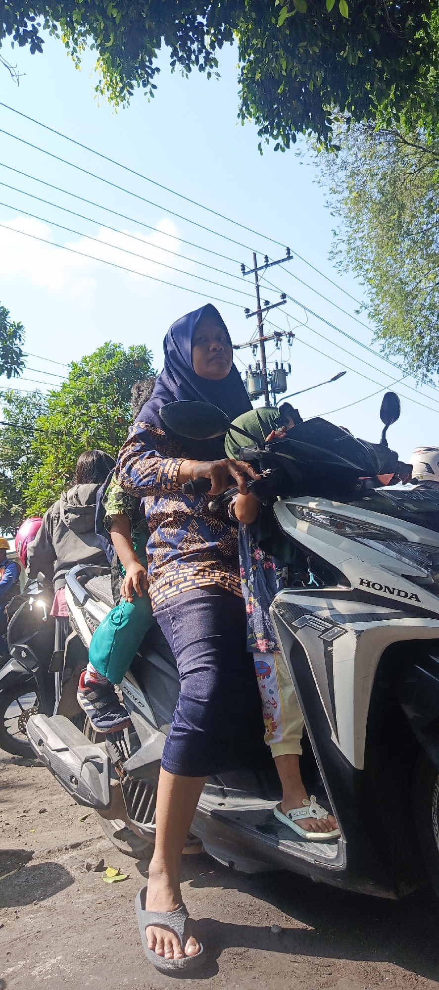 The image shows a woman wearing a blue hijab and a patterned top, sitting on a Honda motorcycle. She is looking forward and appears to be holding the handlebars. To her right, a child wearing a patterned outfit is seated behind her. The child's legs are visible, and they are wearing light-colored sandals with a bunny design. Behind the woman, another person is partially visible, wearing a grey jacket and a pink helmet. The motorcycle is on a dirt road, with some leaves scattered on the ground. In the background, there are trees, electrical poles with transformers, and power lines against a bright blue sky. The scene appears to be in Krian, Indonesia, as indicated by the context.