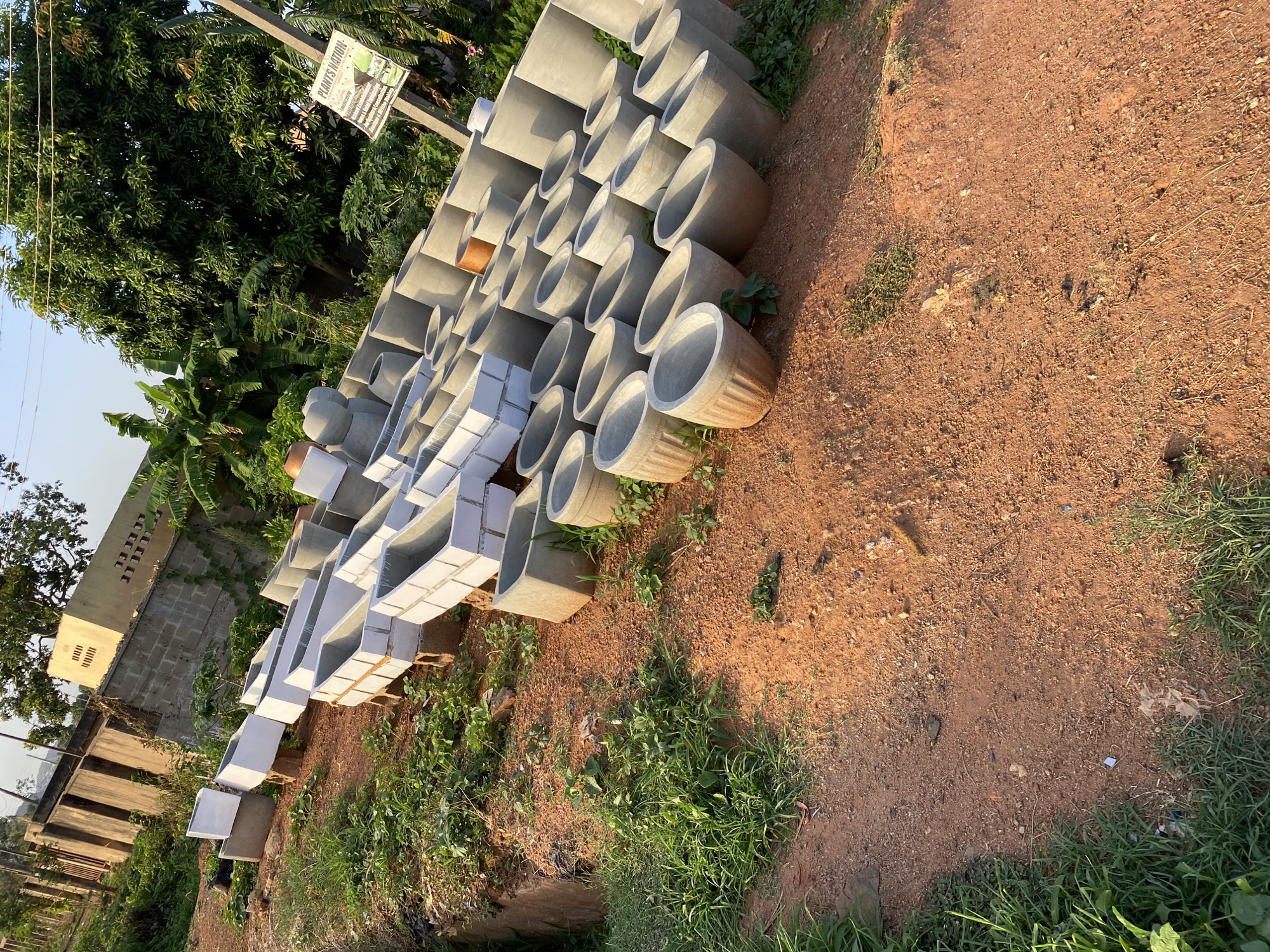 The image shows a collection of concrete cylindrical and rectangular structures, likely planters or construction materials, stacked outdoors on reddish-brown soil mixed with grass and weeds. In the background are trees and foliage, a building, and a signpost with a sign that says, "Plants Nation." The location is identified as Bukuru, Nigeria. No people are visible.