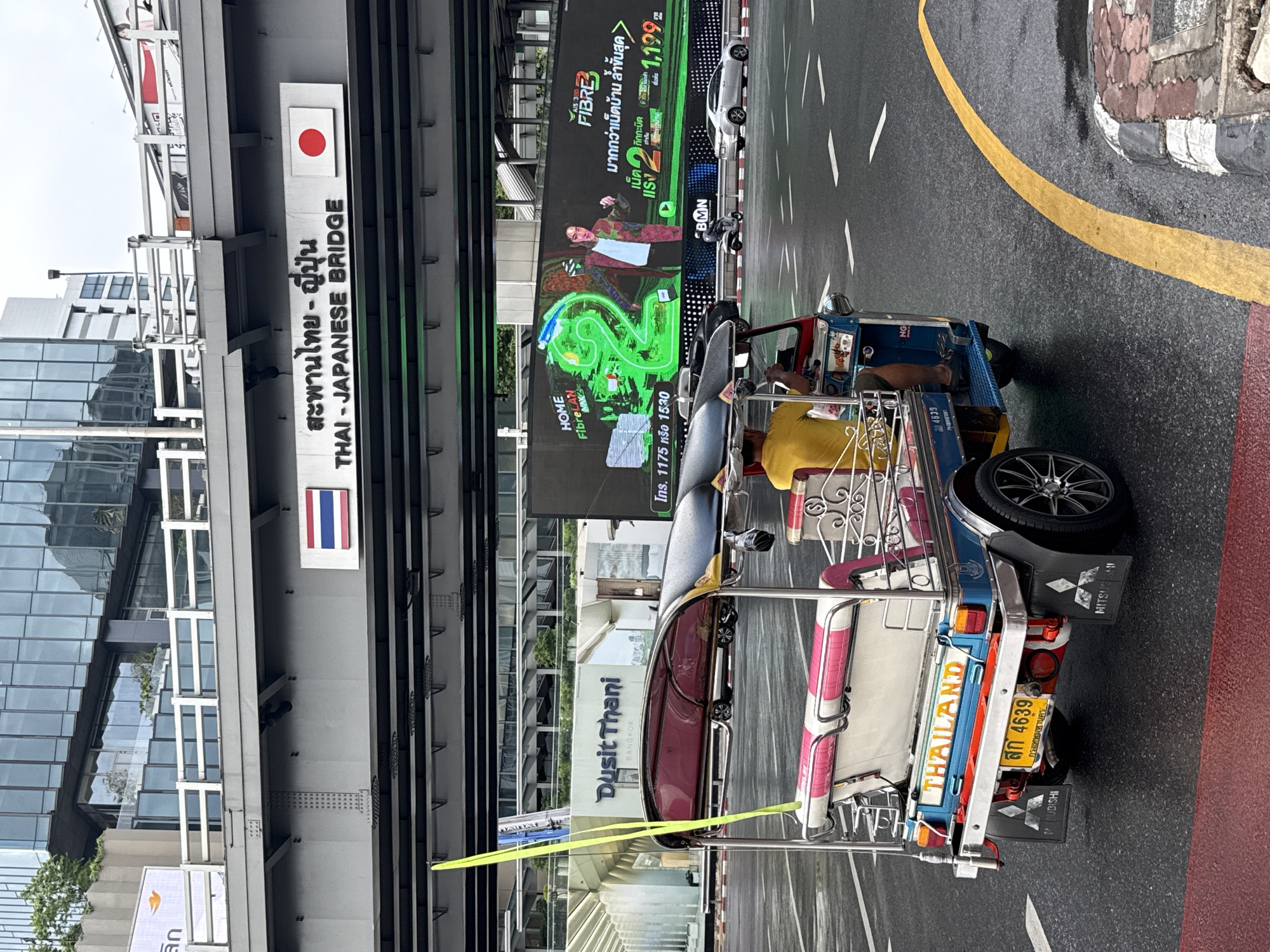 Here is a concise description of the image:
The photo shows a tuk-tuk in Bangkok, Thailand, positioned near the Thai-Japanese Bridge, as indicated by a sign with both Thai and English lettering, as well as the flags of Thailand and Japan. The tuk-tuk has a yellow license plate that reads "สก 4639," indicating it is registered in Bangkok. The rear of the tuk-tuk displays "THAILAND". Nearby, an advertisement for "Fibre" is displayed on a large digital screen. A building with the name "Dusit Thani Bangkok" is visible, suggesting a hotel or business in the vicinity.