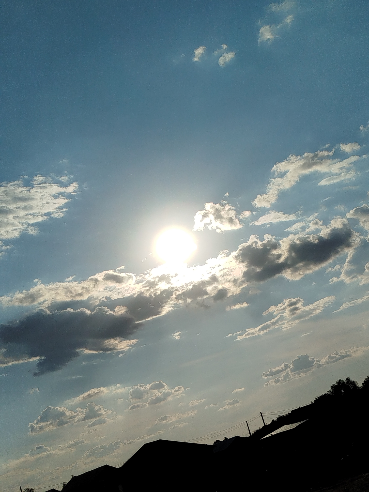 The image shows a sky with a bright sun partially obscured by clouds. The sun is located in the upper center of the frame, with rays emanating outwards. Various cloud formations are present, ranging from thin wispy clouds to darker, more defined cumulus clouds. The bottom of the image is silhouetted against the sky, showing the dark shapes of rooftops and trees, suggesting an outdoor scene in a residential or semi-rural area. There are power lines visible in the foreground. The lighting suggests it is either sunrise or sunset, with the sun low in the sky.