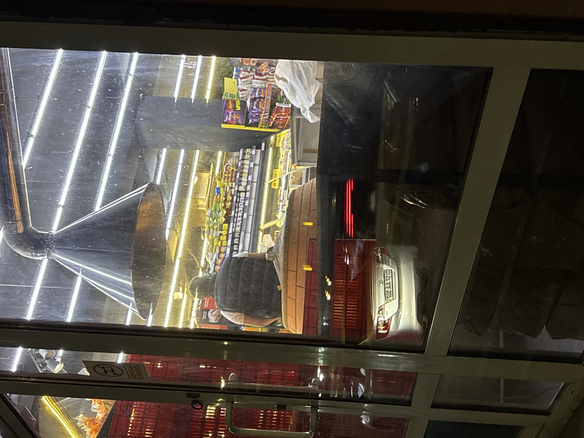 The image, rotated 90 degrees clockwise from its natural orientation, depicts a view through a glass window or door into a brightly lit store, overlaid with strong reflections from the outside.
Inside the store, a person is seen from behind, wearing a dark puffer vest over a lighter shirt, crouching or bending down near shelves stocked with various packaged goods, including chips and snacks. The store's interior is illuminated by numerous bright, cool-toned LED strip lights on the ceiling. A large, metallic ventilation pipe or duct is visible on the left side of the store's ceiling.
Significant reflections on the glass surface show elements from the exterior. Most notably, the rear of a white car is prominently reflected on the right. Its license plate, "30 AA 345", is clearly visible and is characteristic of vehicles in Armenia. The dark appearance of the reflected exterior suggests it is nighttime. Red light streaks, possibly from the car's taillights or other external sources, are also reflected.
In the lower-left corner of the glass, a "No Smoking" sign is visible, featuring a red circle with a crossed-out cigarette. Below it, Armenian text reads "ՏՐԱՄԱԴՐՈՒՄ Է 50000 ԴՐԱՄ", which translates to "PROVIDES 50,000 DRAM", indicating a fine. Red plastic crates are also faintly reflected at the bottom. The scene likely takes place at night in a commercial area of Yerevan, Armenia.